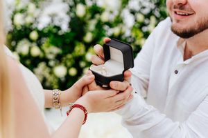 Man in white shirt presenting an open ring box during a romantic marriage proposal outdoors with white flowers.
