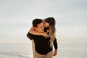 A couple embraces joyfully on a beach, the woman hugging the man from behind as ocean waves shimmer in the background.