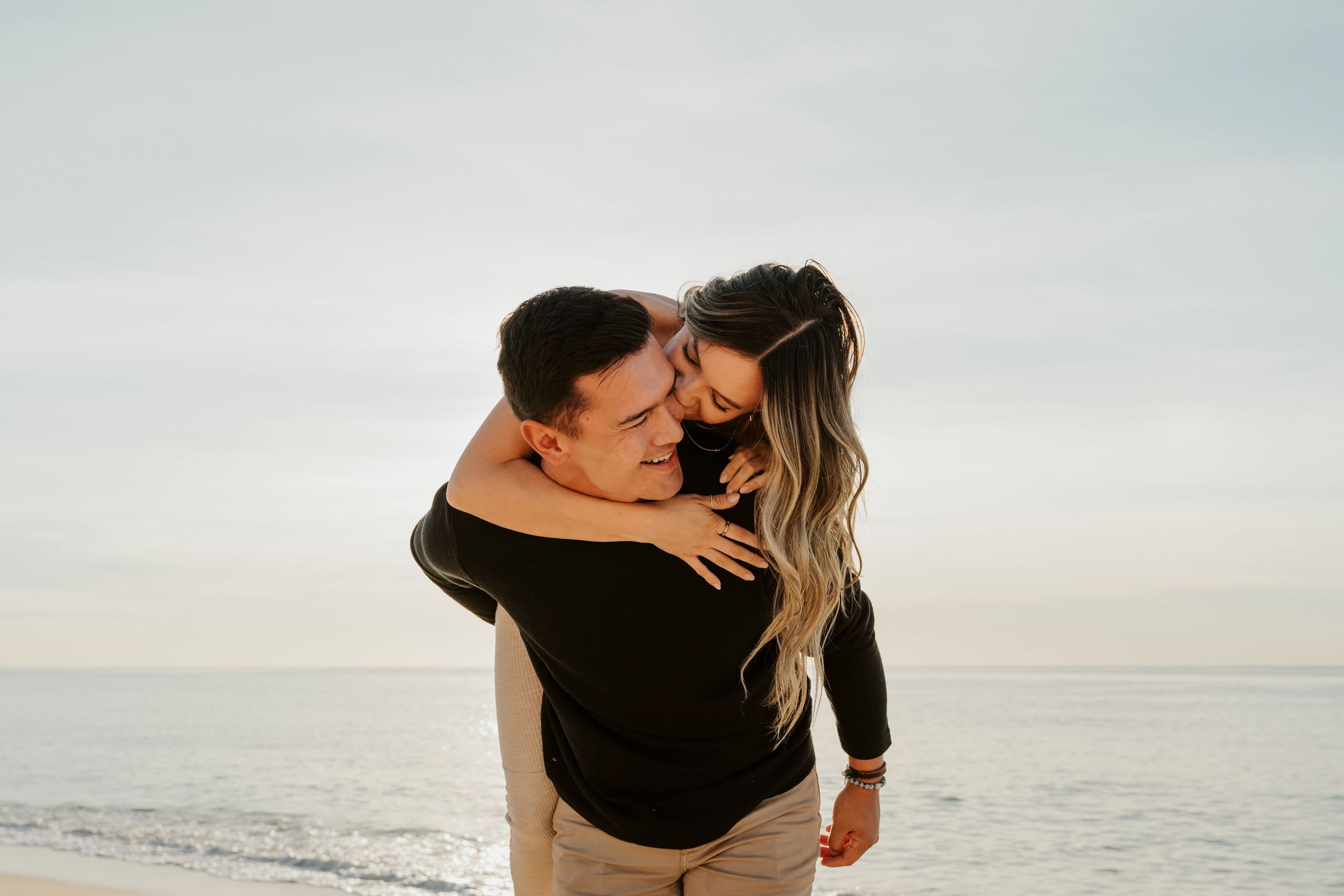A couple embraces joyfully on a beach, the woman hugging the man from behind as ocean waves shimmer in the background.