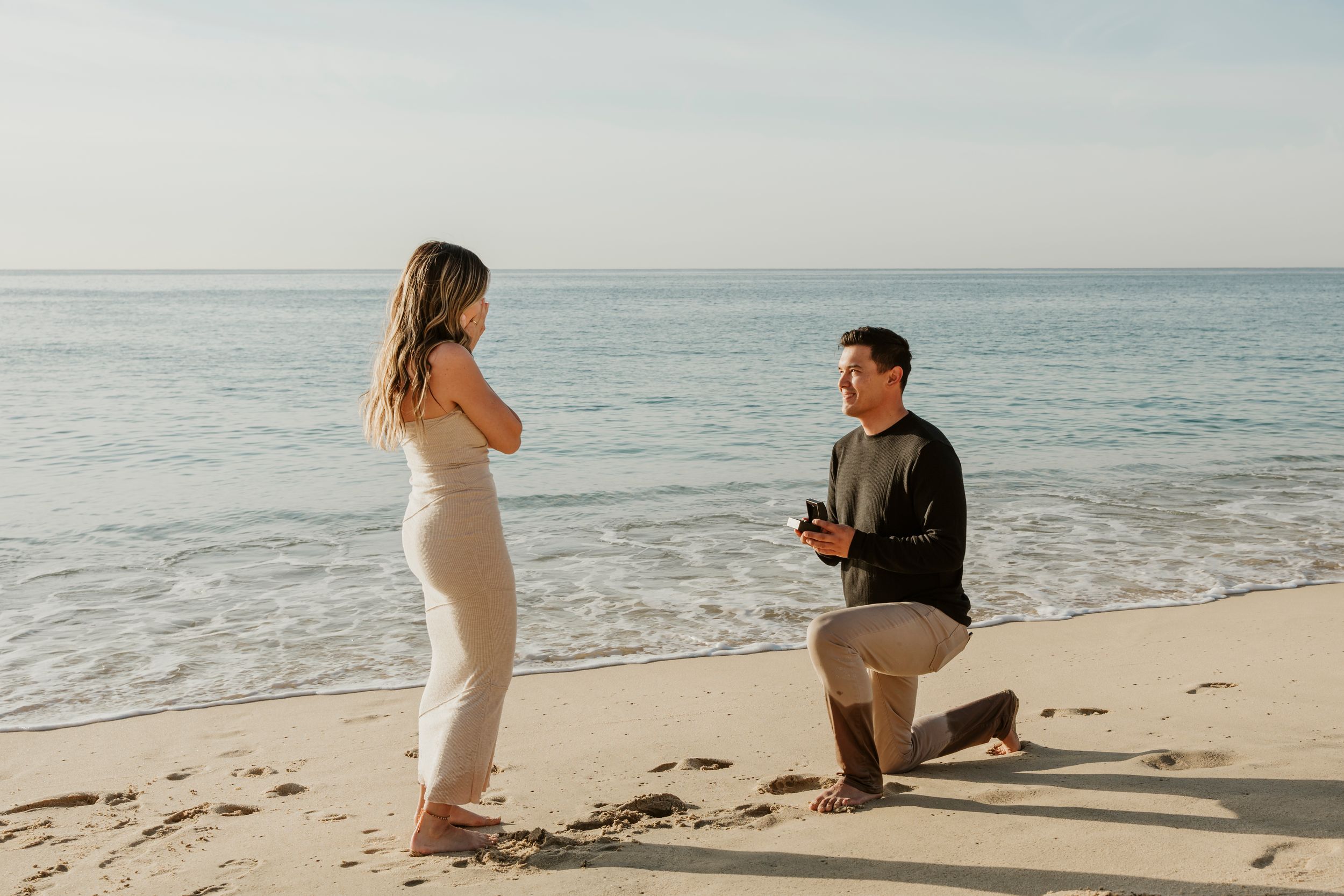 Man proposing on one knee to woman on sandy beach with ocean backdrop in romantic sunset setting.