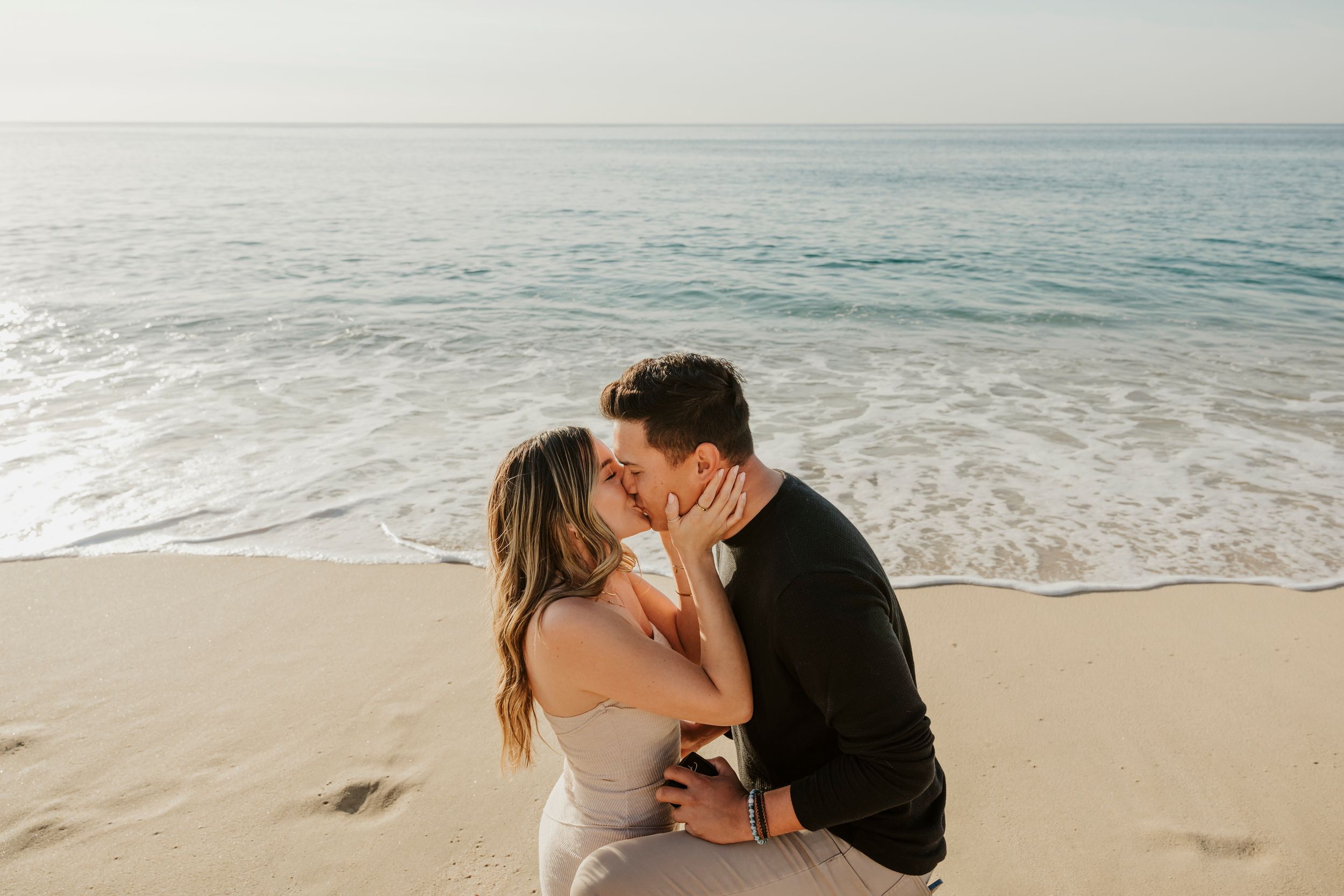 Couple sharing a romantic kiss on a sandy beach with calm ocean waves in the background.