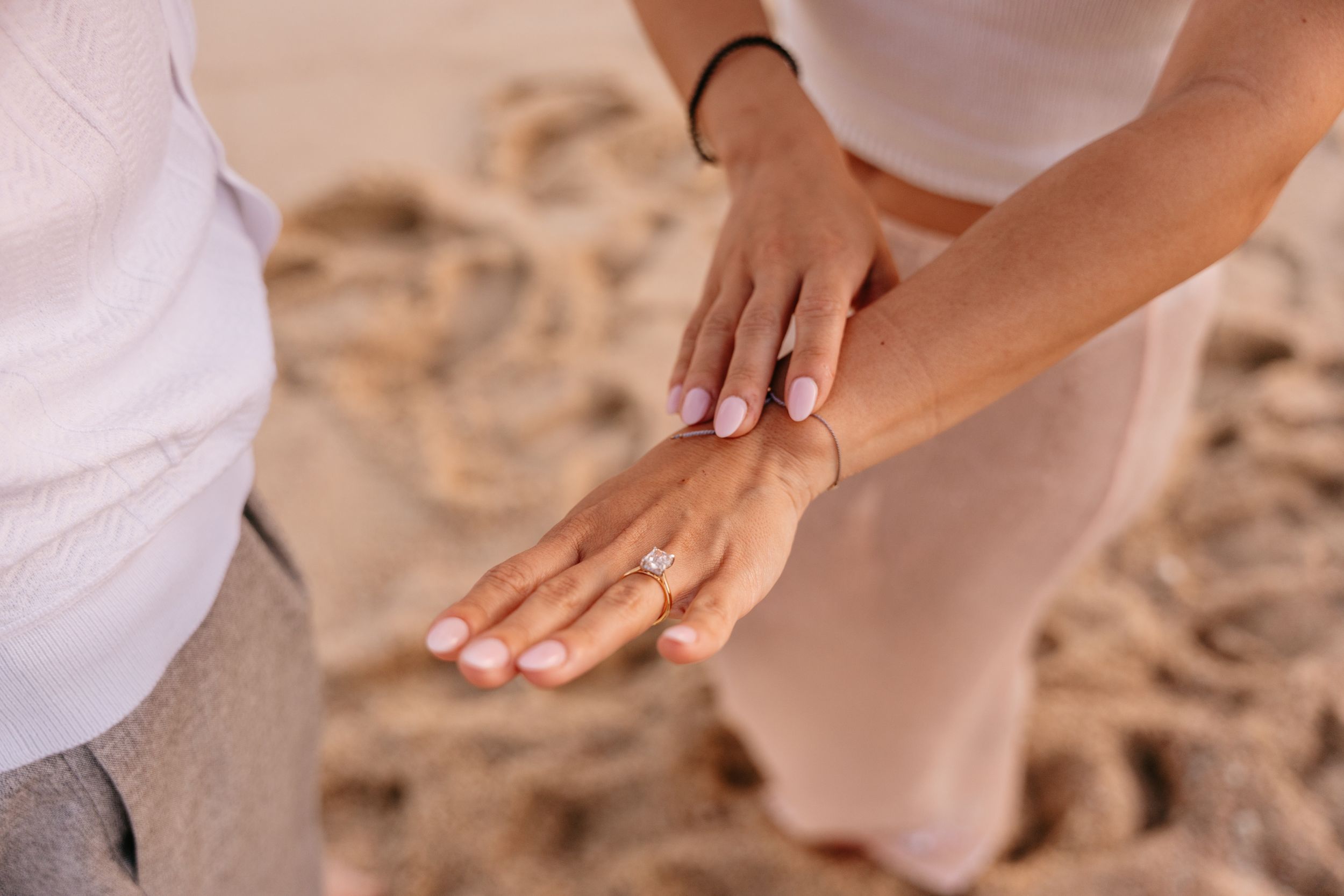 Close-up of couple's hands on sandy beach, woman wearing diamond engagement ring, romantic proposal moment.