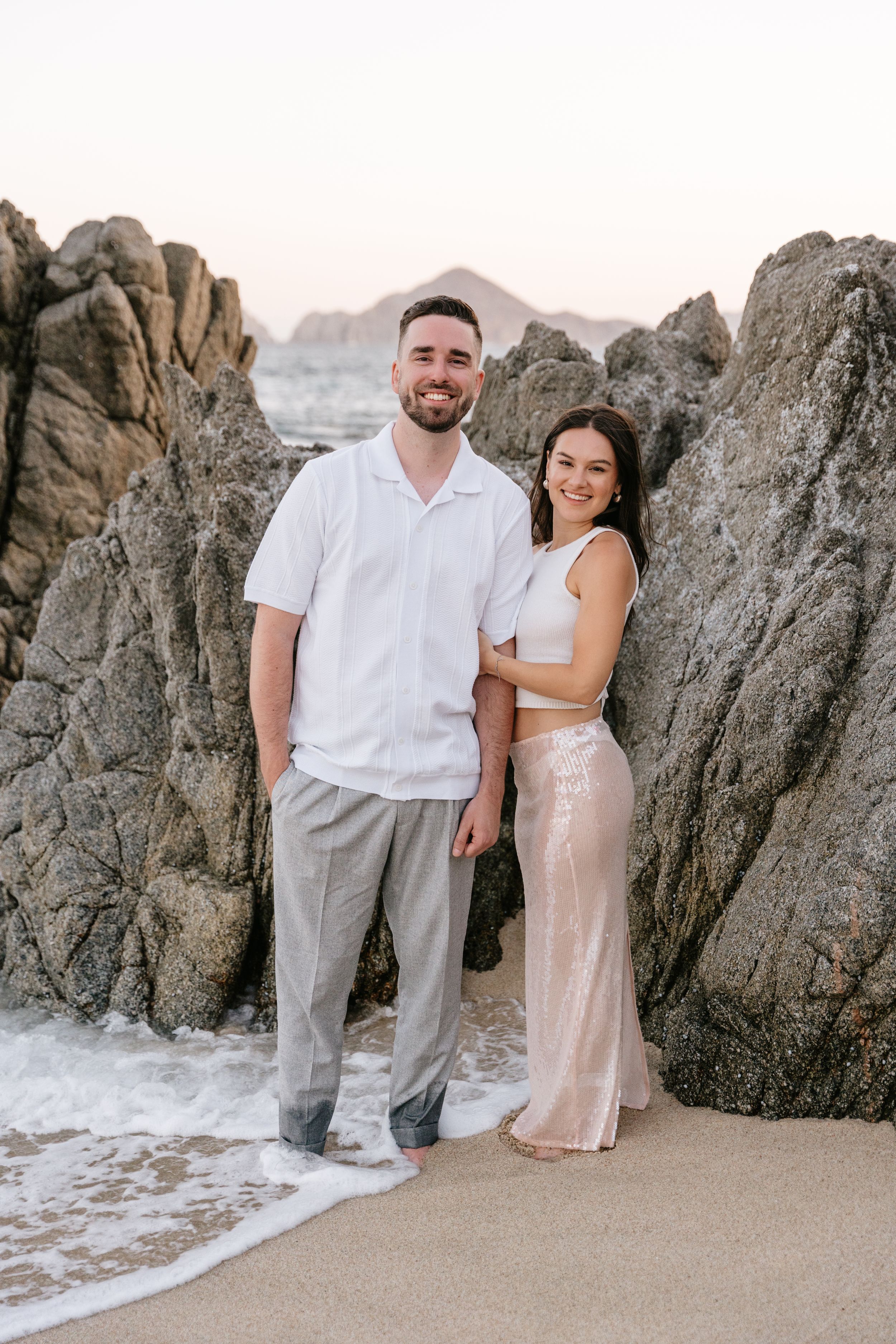 Couple posing on a sandy beach surrounded by large rocks at sunset, man in white shirt and woman in sparkly skirt.