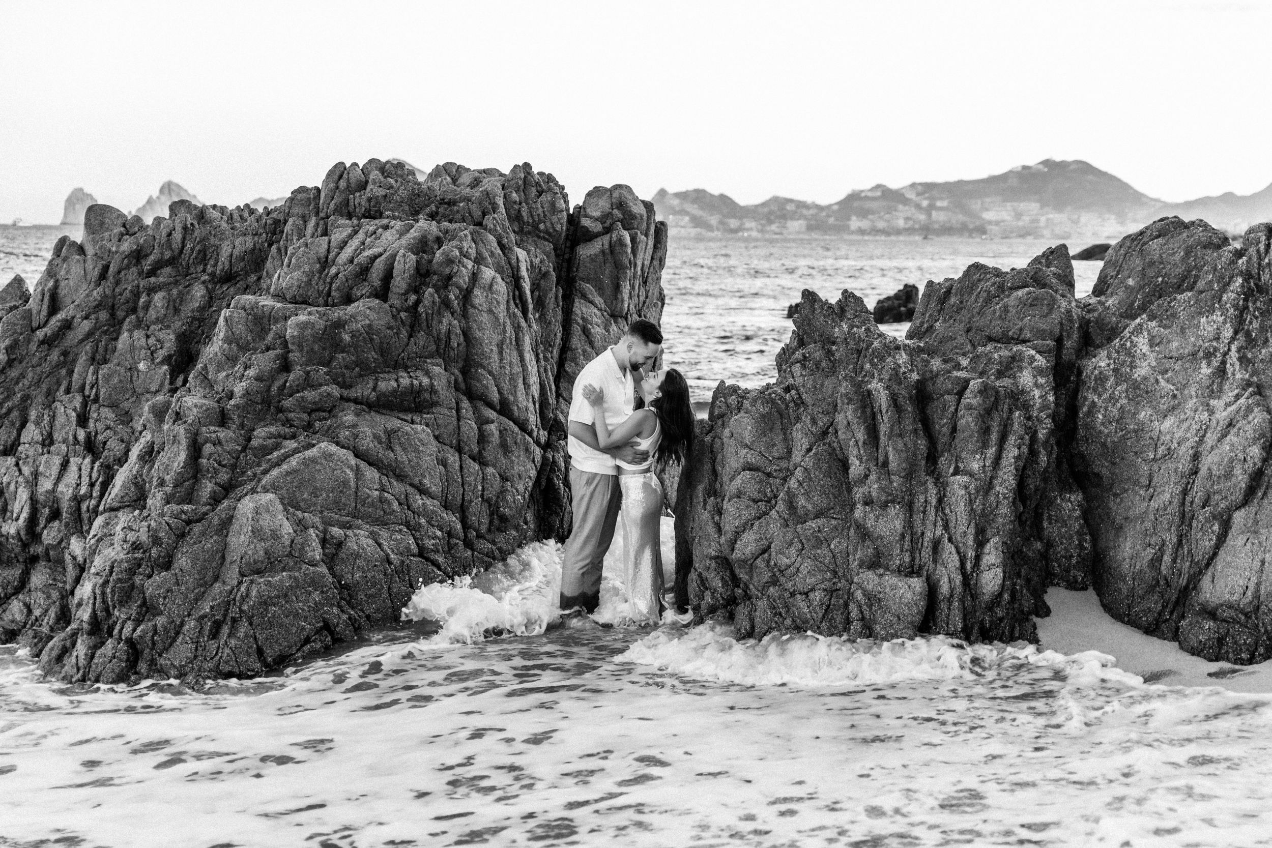 Couple embracing between rocky coastal formations with ocean waves, black and white beach photography.