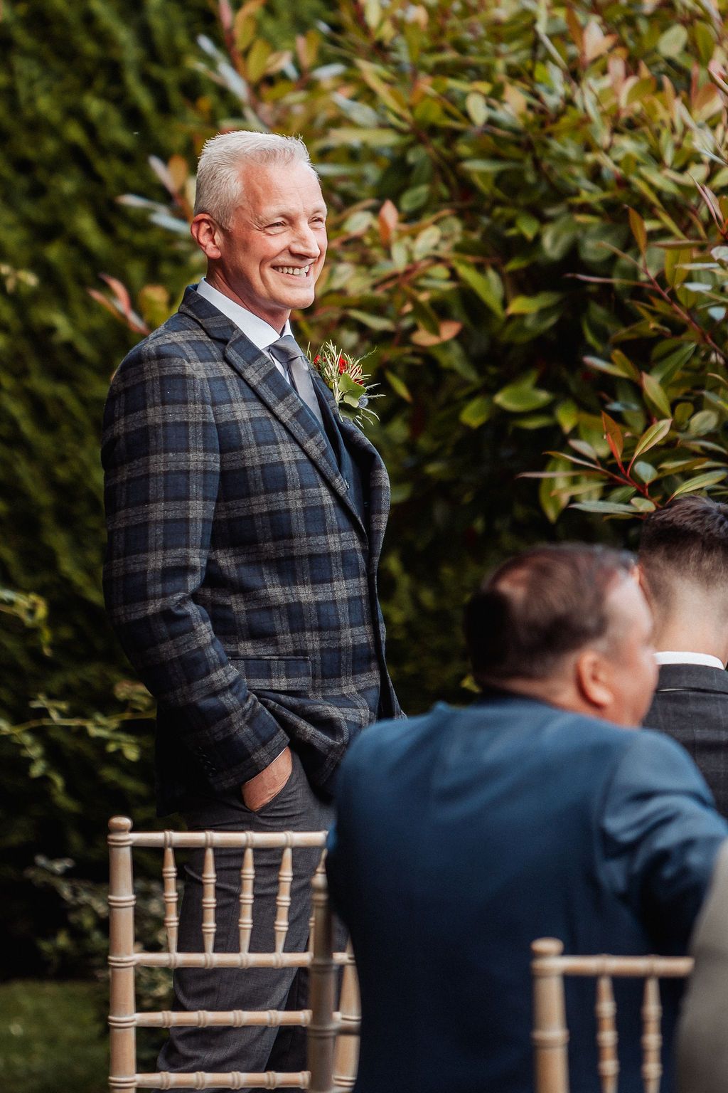Smiling older man in plaid suit with boutonniere stands at outdoor wedding ceremony surrounded by greenery.