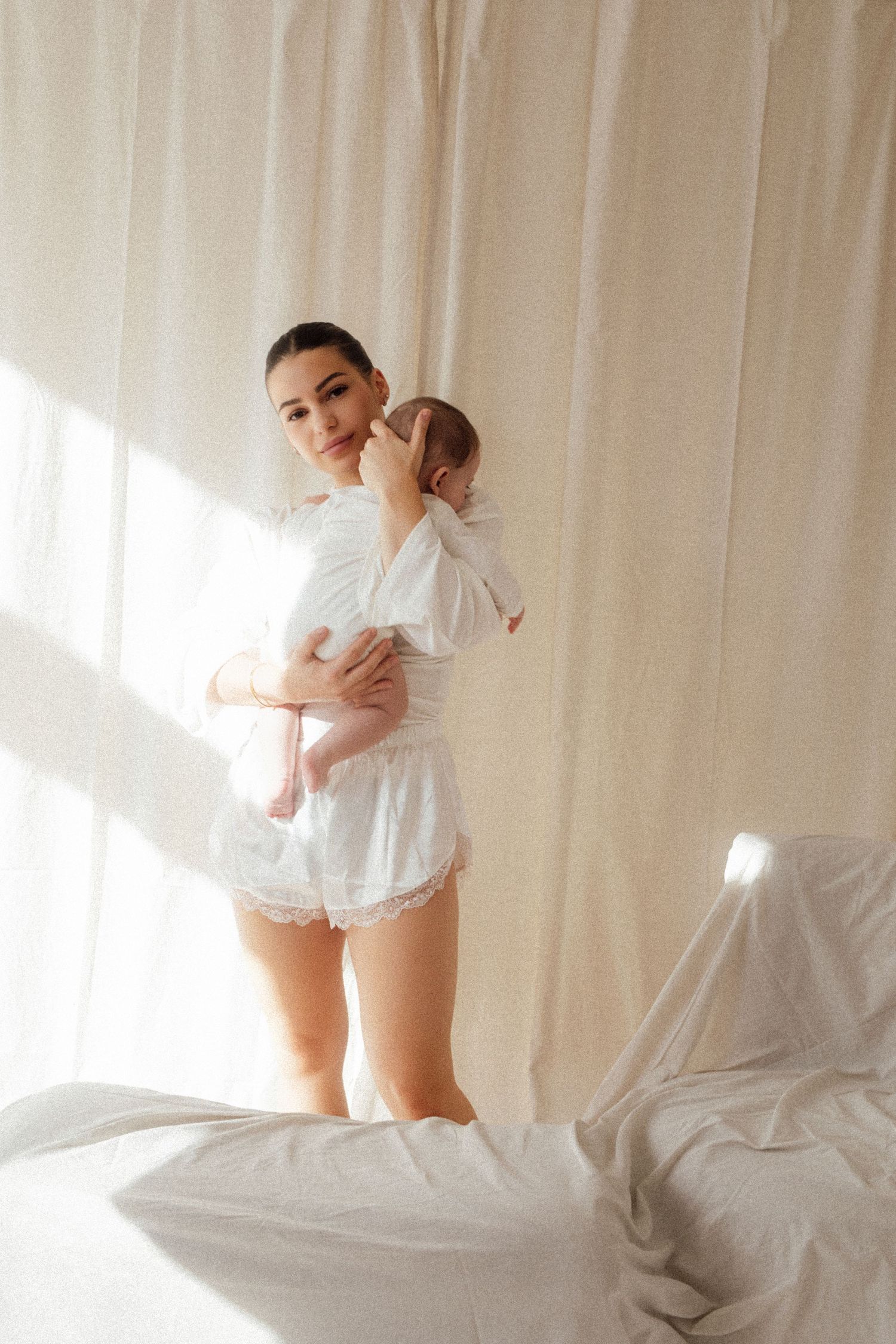 Young mother in white lace shorts set holds newborn baby near bright curtained window.