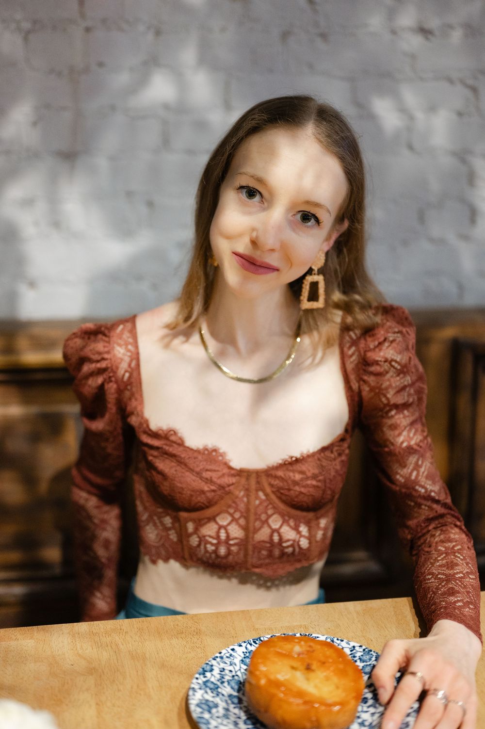 Young woman in brown lace top with gold earrings sitting at cafe table with pastry on blue plate.