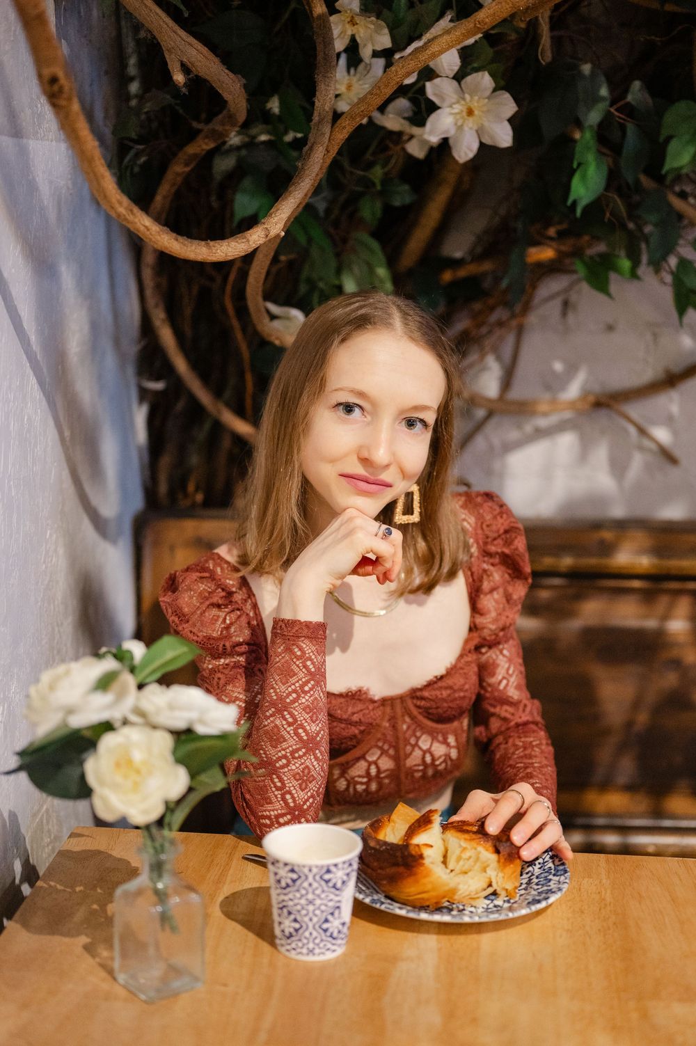 Young woman in brown lace top enjoying coffee and pastry at a cozy cafe table with white flowers.