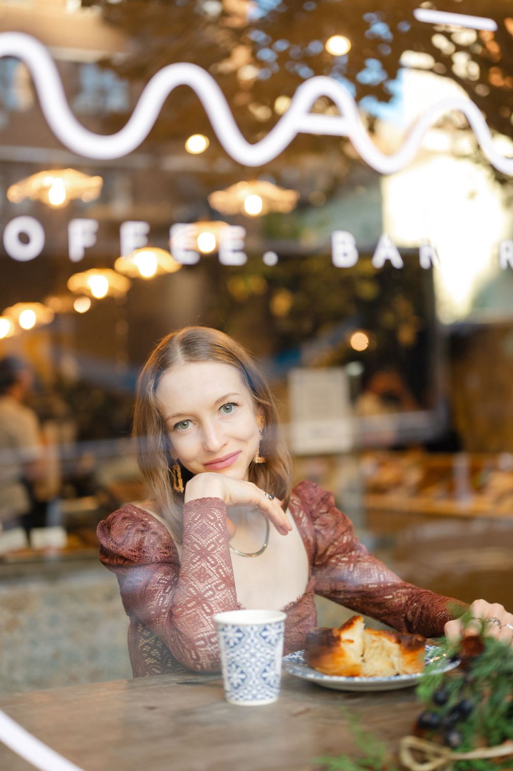 Woman smiling at camera in cozy coffee bar, sitting at table with cup and pastry, wearing lace top.