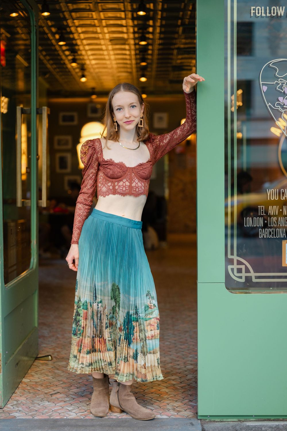 Woman in lace crop top and teal floral pleated midi skirt with ankle boots posing outside a restaurant.