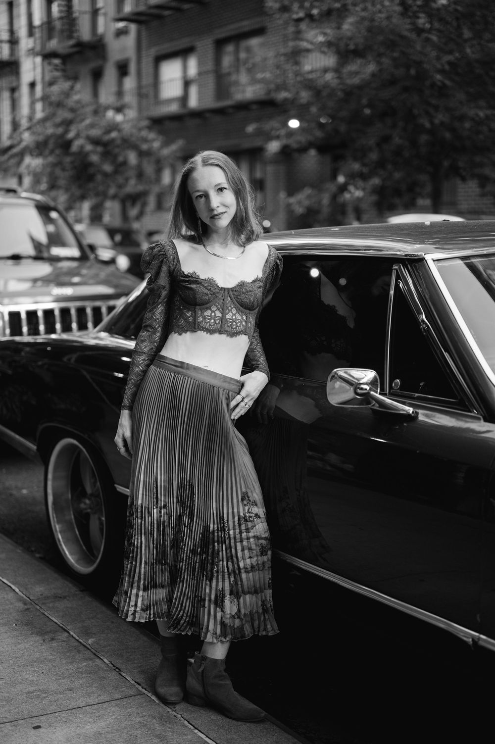 Black and white photo of woman in lace crop top and pleated skirt leaning against a classic car.