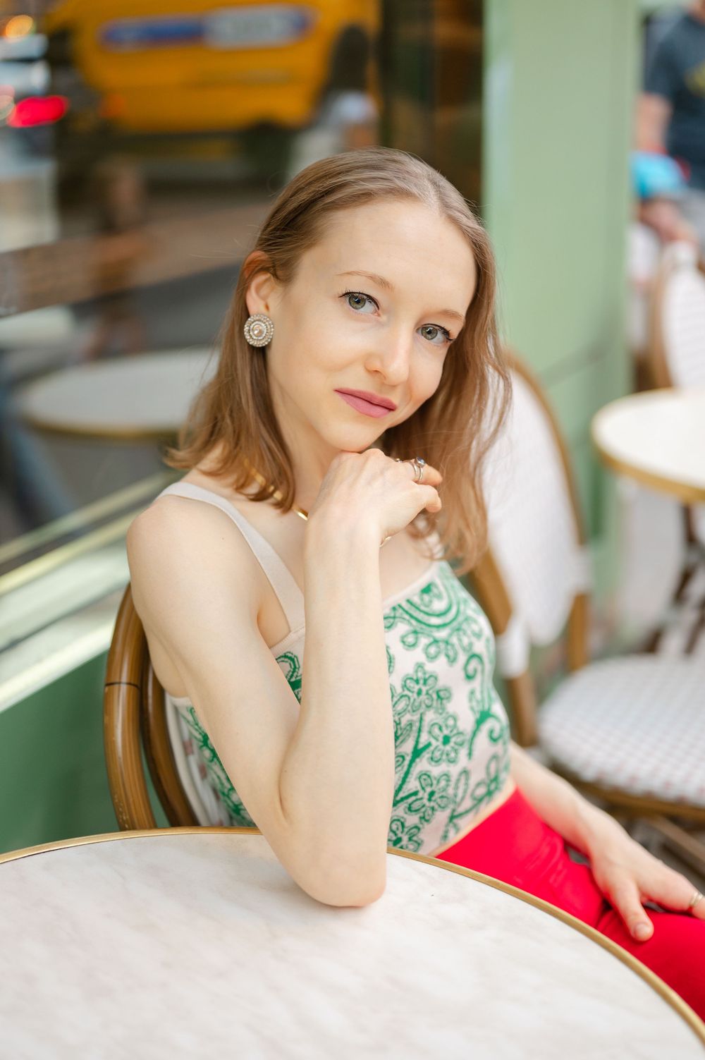 Young woman in green floral top and red pants sitting at outdoor cafe table wearing stud earrings.
