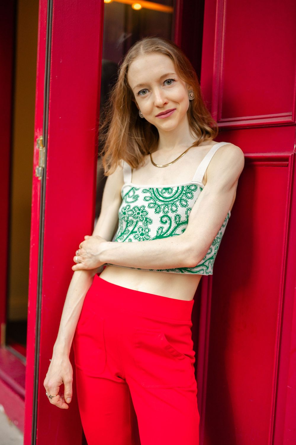 A slender woman in a green embroidered crop top and red pants poses against a vibrant red door.