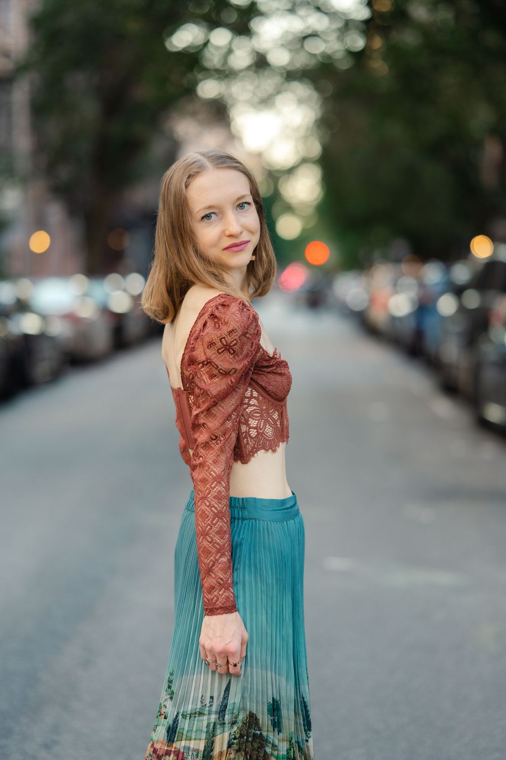 Young woman in rust lace crop top and teal pleated skirt posing on a blurred city street at dusk.