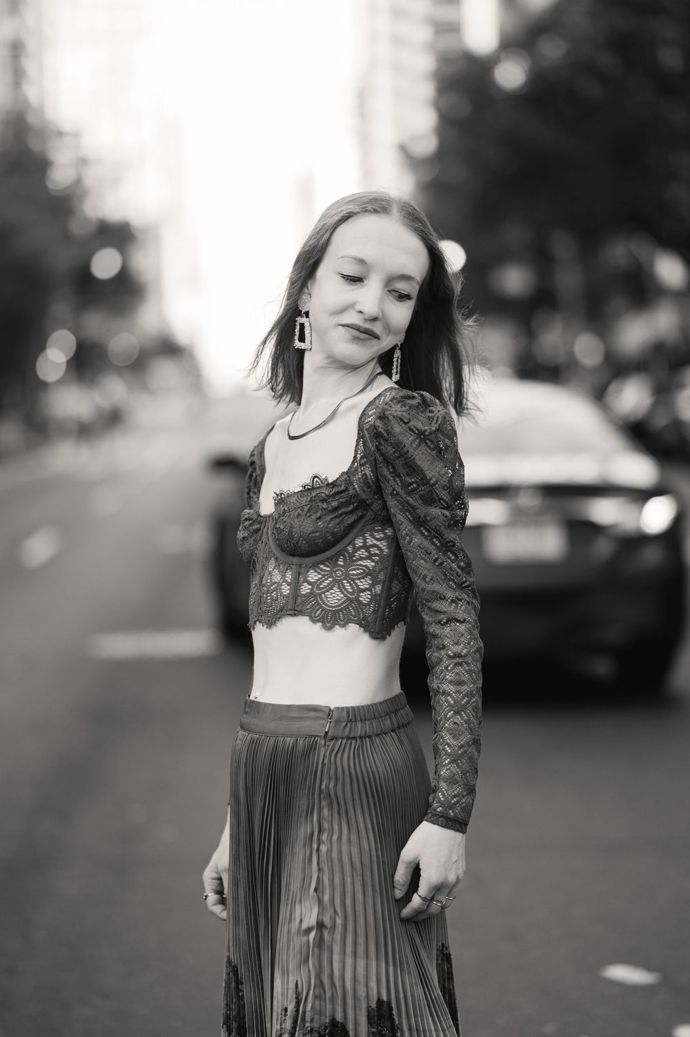 Young woman in lace crop top and pleated skirt posing on a city street in black and white photo.
