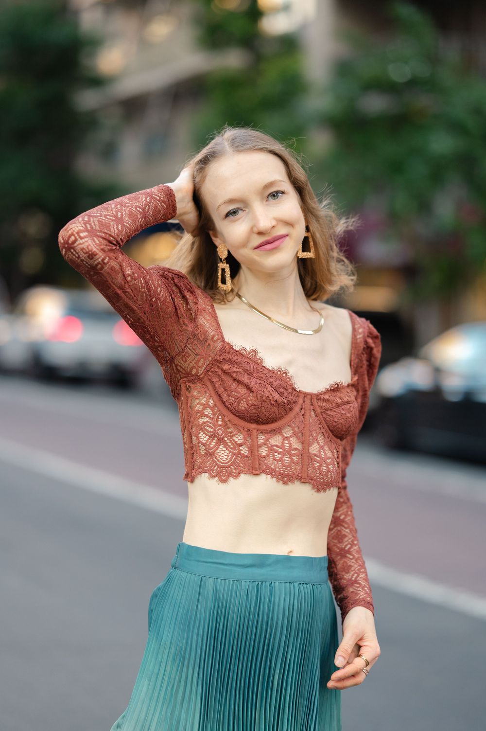 Stylish woman in brown lace bustier top and teal pleated skirt posing outdoors on a city street.