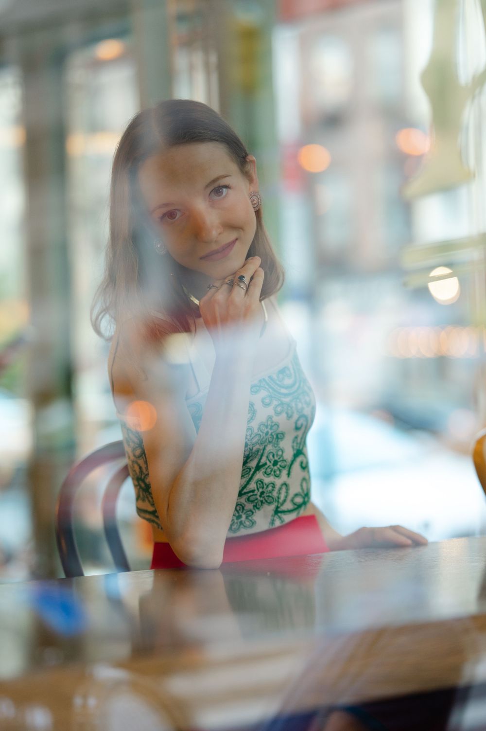 Woman in green patterned top sitting at a cafe table photographed through a reflective window.