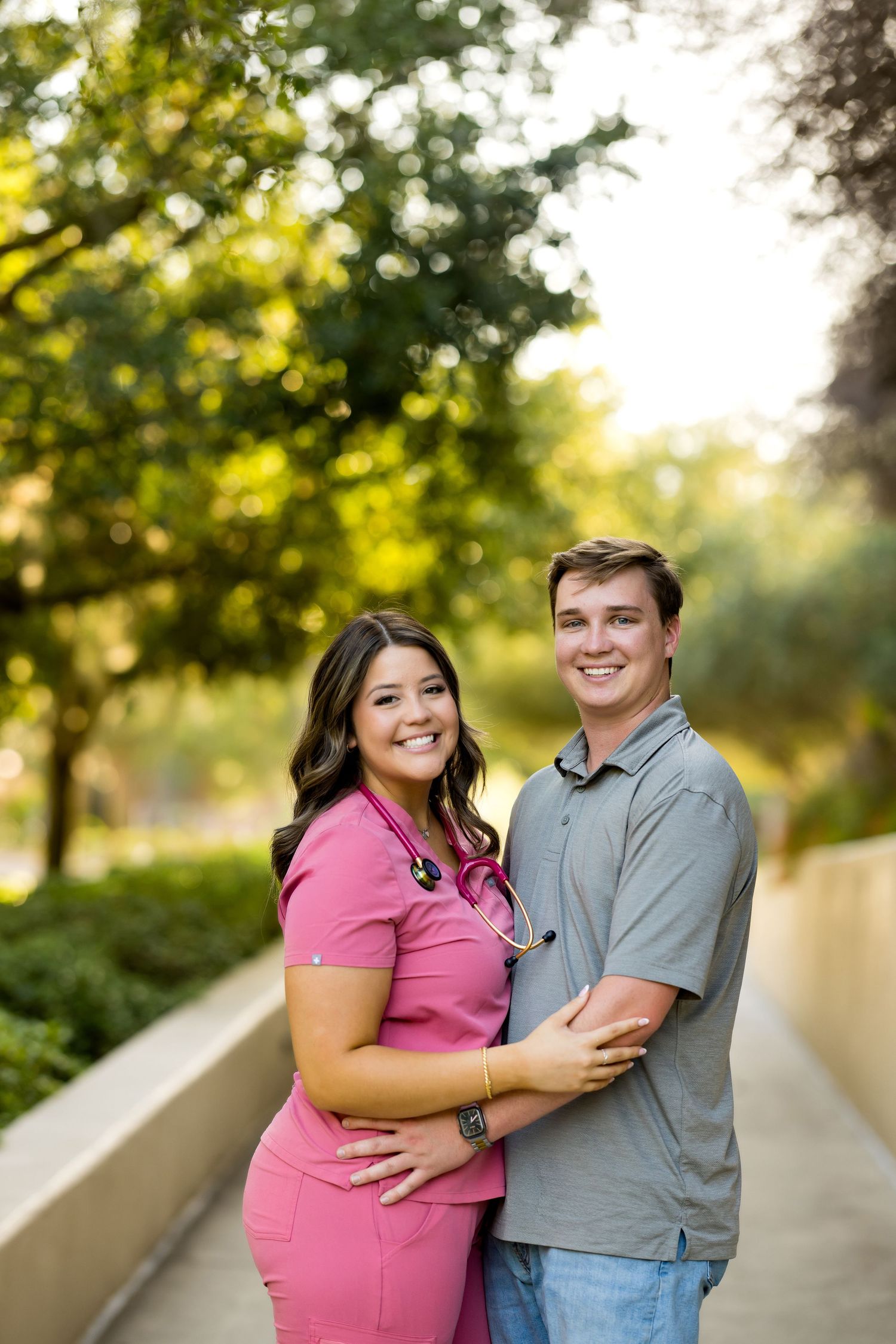 A smiling couple poses outdoors, the woman wearing pink nursing scrubs with a stethoscope, embraced by a man in a gray shirt.