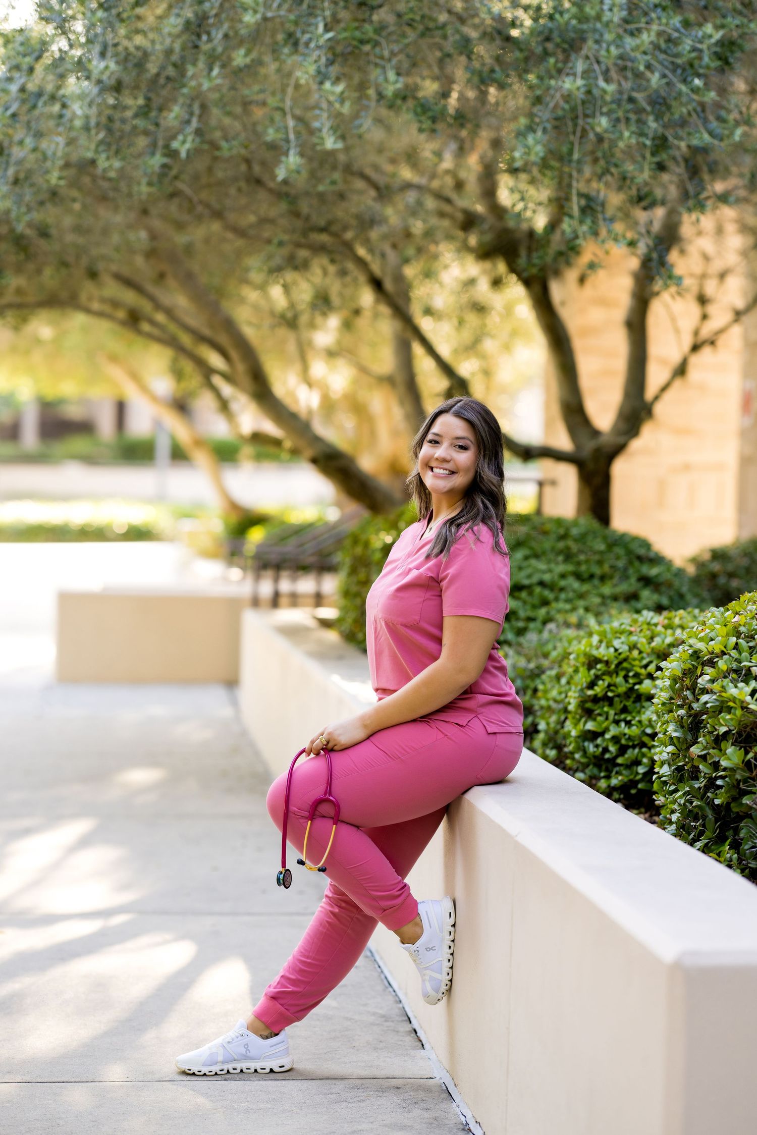 Young woman in pink scrubs with stethoscope sitting on outdoor wall, smiling, surrounded by lush green trees.