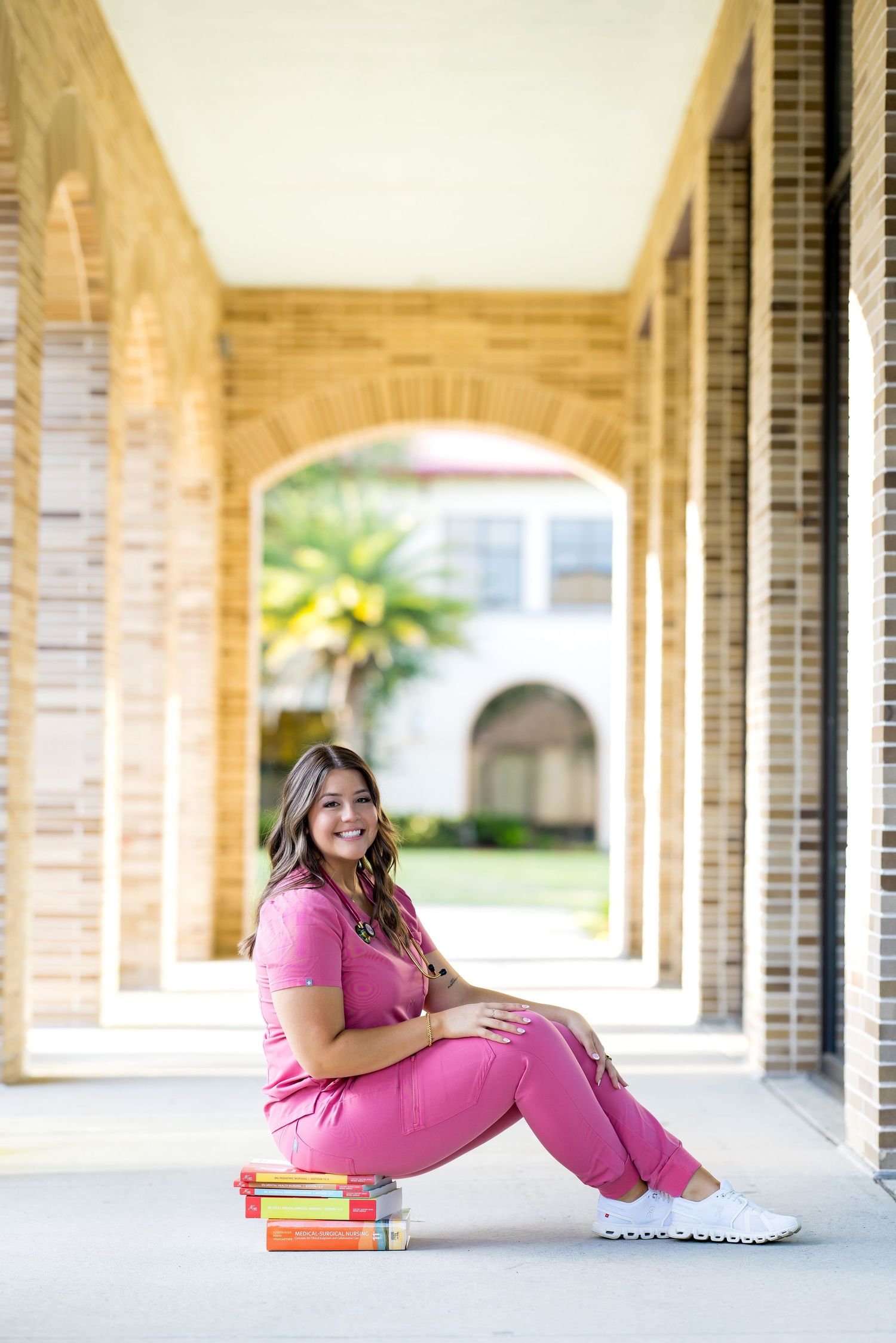 Young woman in pink scrubs sits on books in a brick archway corridor, smiling for a nursing graduation photo.