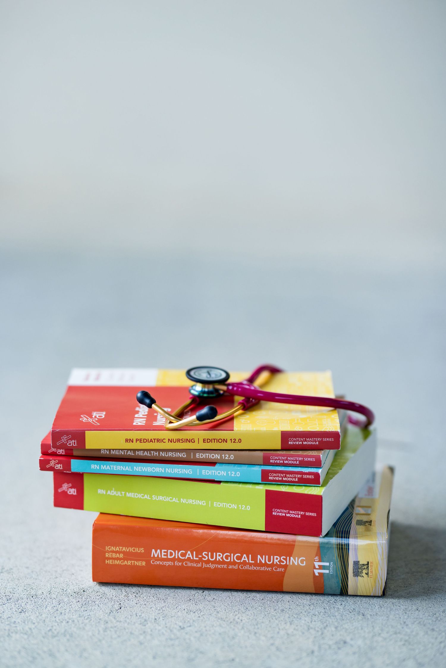 Stack of colorful nursing textbooks with stethoscope on gray background, including Medical-Surgical Nursing edition.