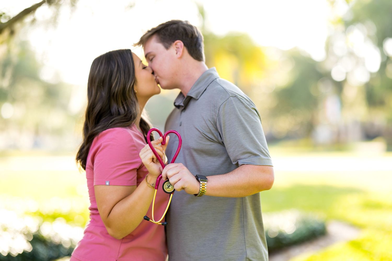 Couple kissing outdoors in a sunny park while holding red heart-shaped props, romantic engagement photo.