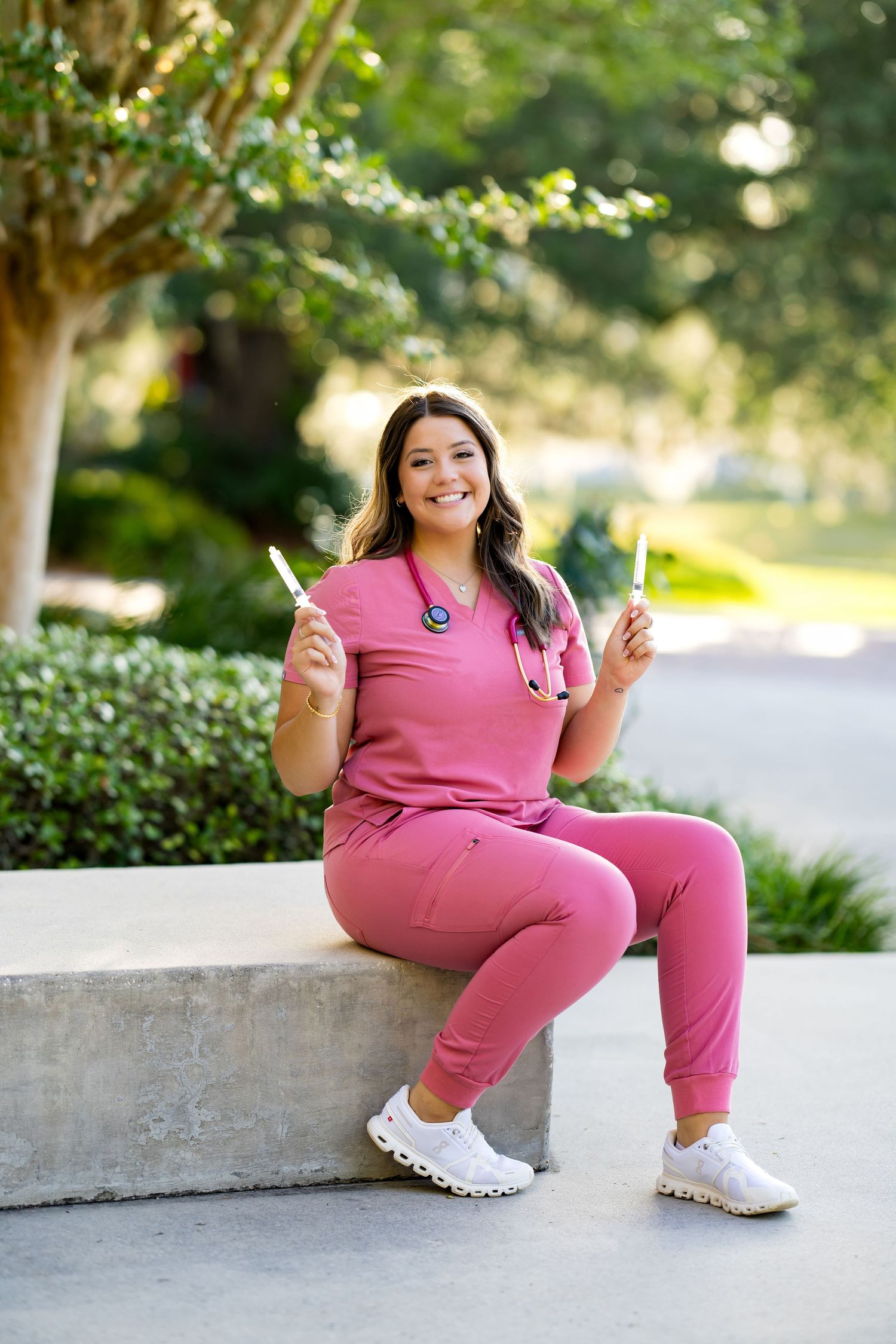 Smiling nurse in pink scrubs sitting outdoors holding medical tools, with stethoscope and white sneakers.