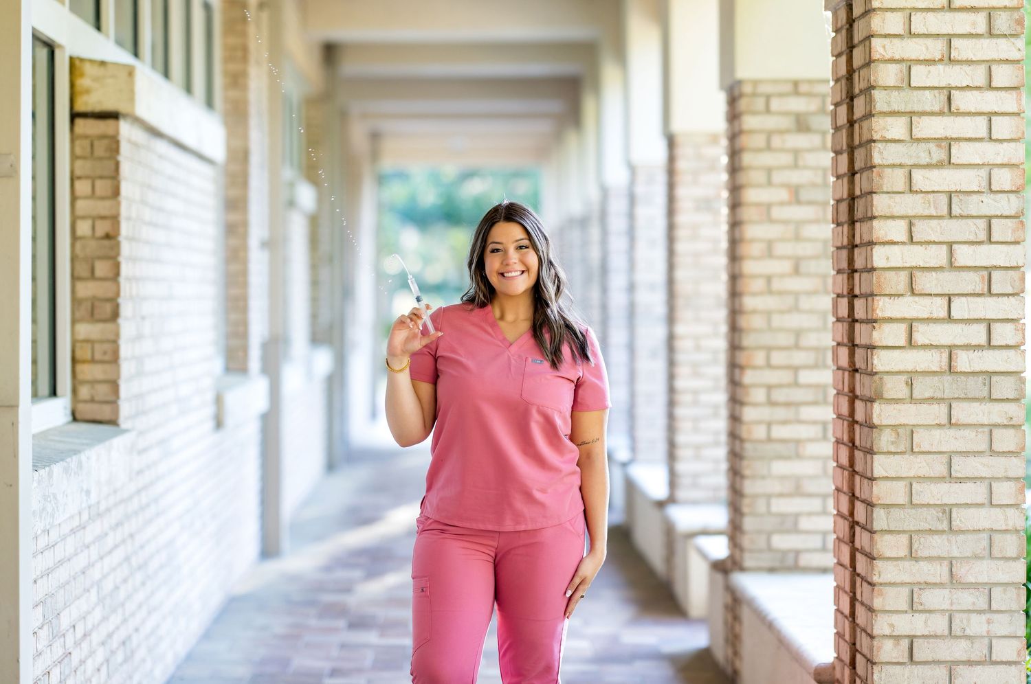 Woman in pink scrubs smiling in a brick hallway, holding a pen, ideal for healthcare professional apparel.