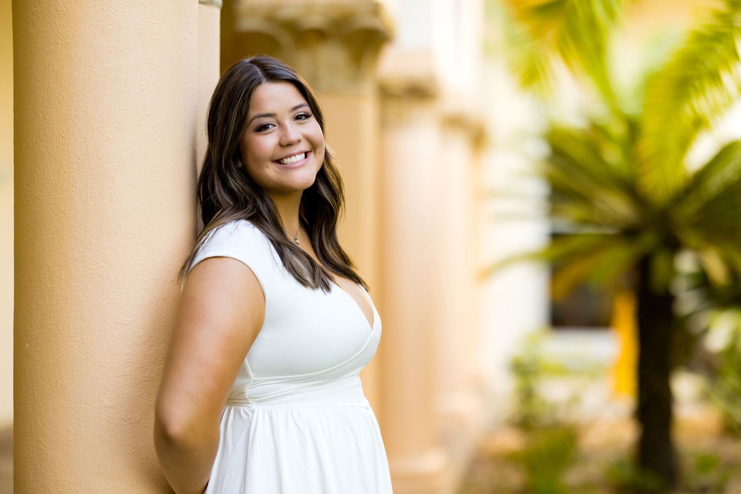 Young woman in white dress smiles leaning against a column surrounded by tropical palm trees outdoors.