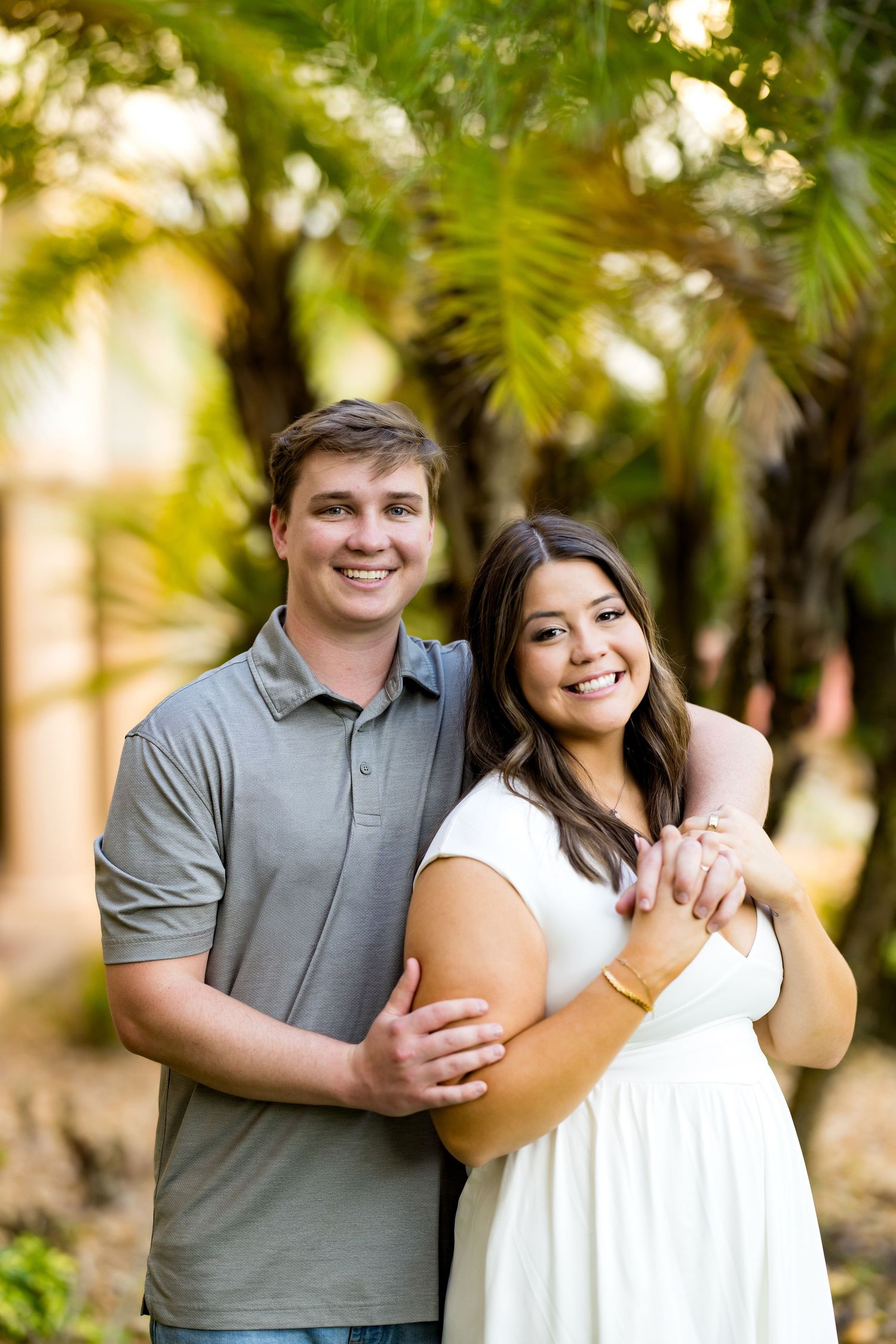 A smiling couple embraces outdoors among lush palm trees, the man in a gray polo and woman in a white dress.