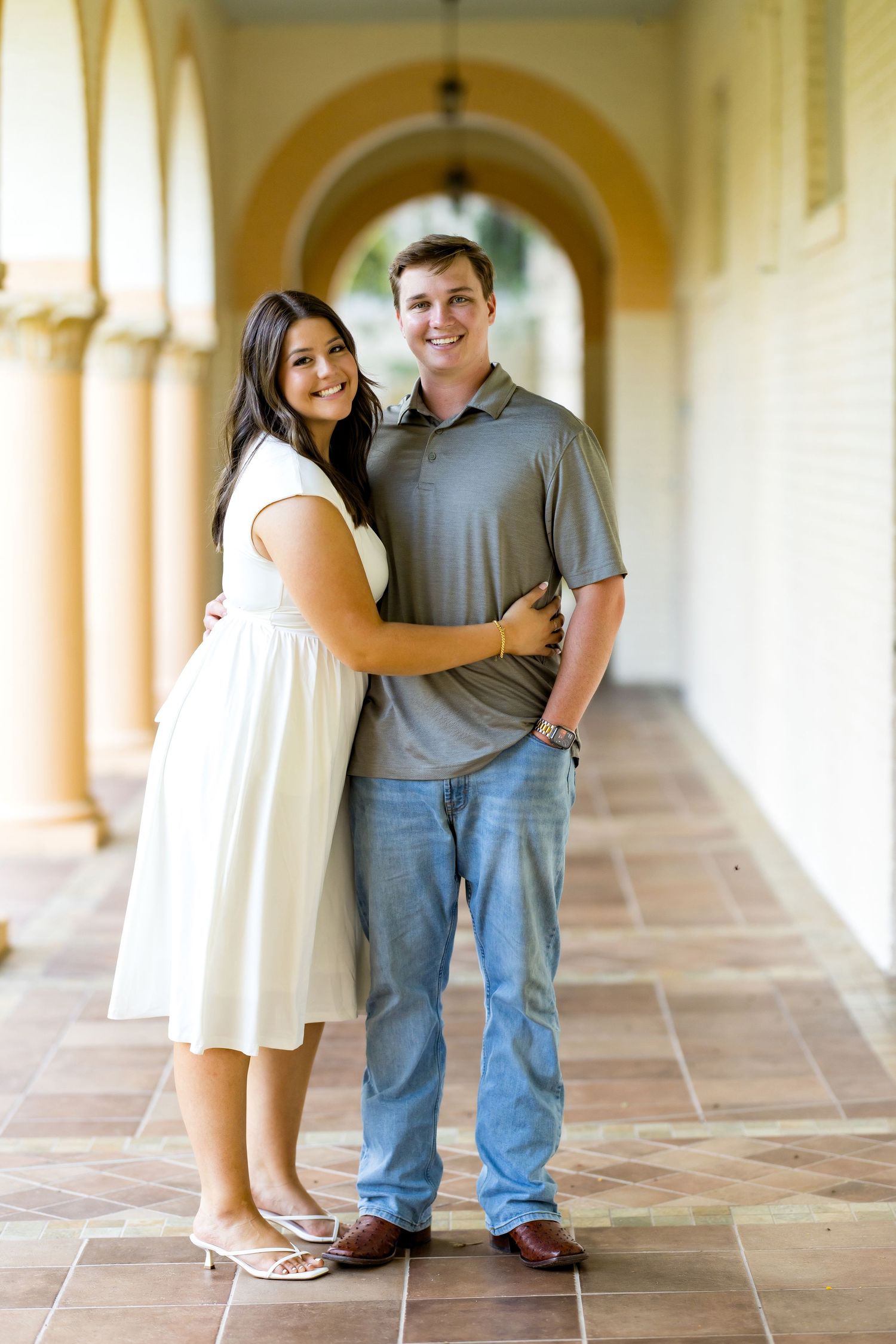 A smiling couple embraces in an arched outdoor corridor, woman in white dress, man in gray polo and jeans.