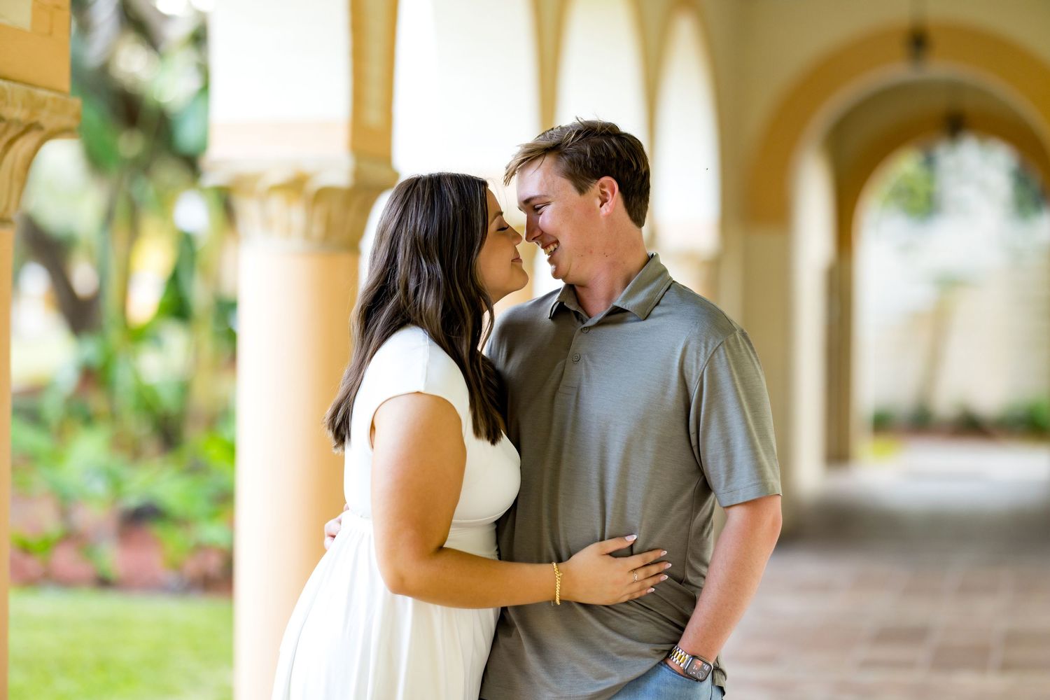 Couple sharing a romantic moment under arched colonnade, woman in white dress and man in gray polo shirt.