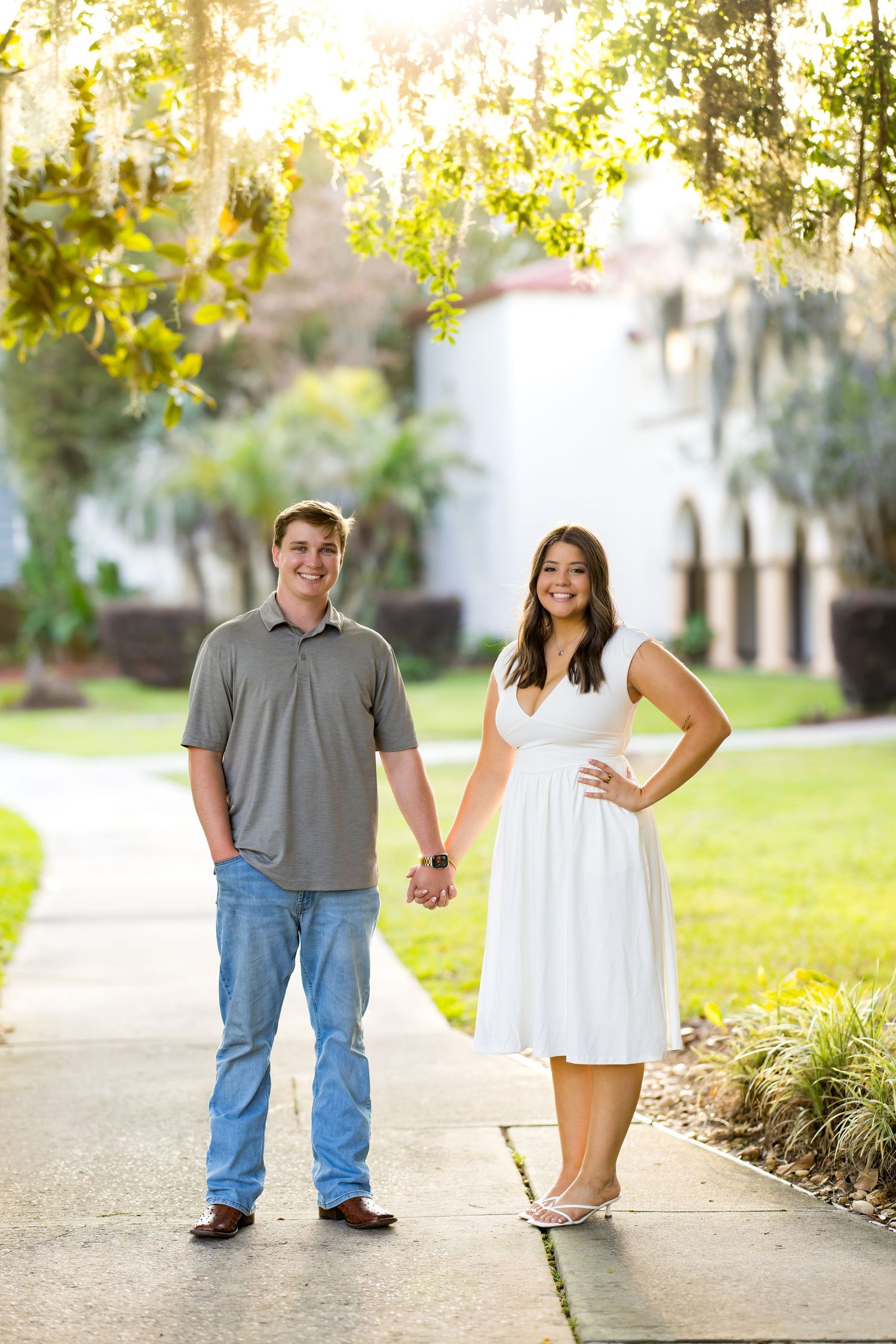 Young couple holding hands on a sunny garden pathway, man in gray polo and jeans, woman in white dress.
