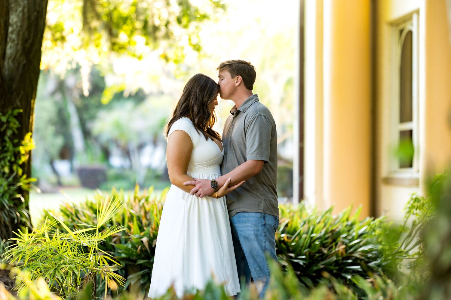 Couple sharing a romantic kiss outdoors among lush tropical greenery, woman in white dress, man in gray shirt and jeans.