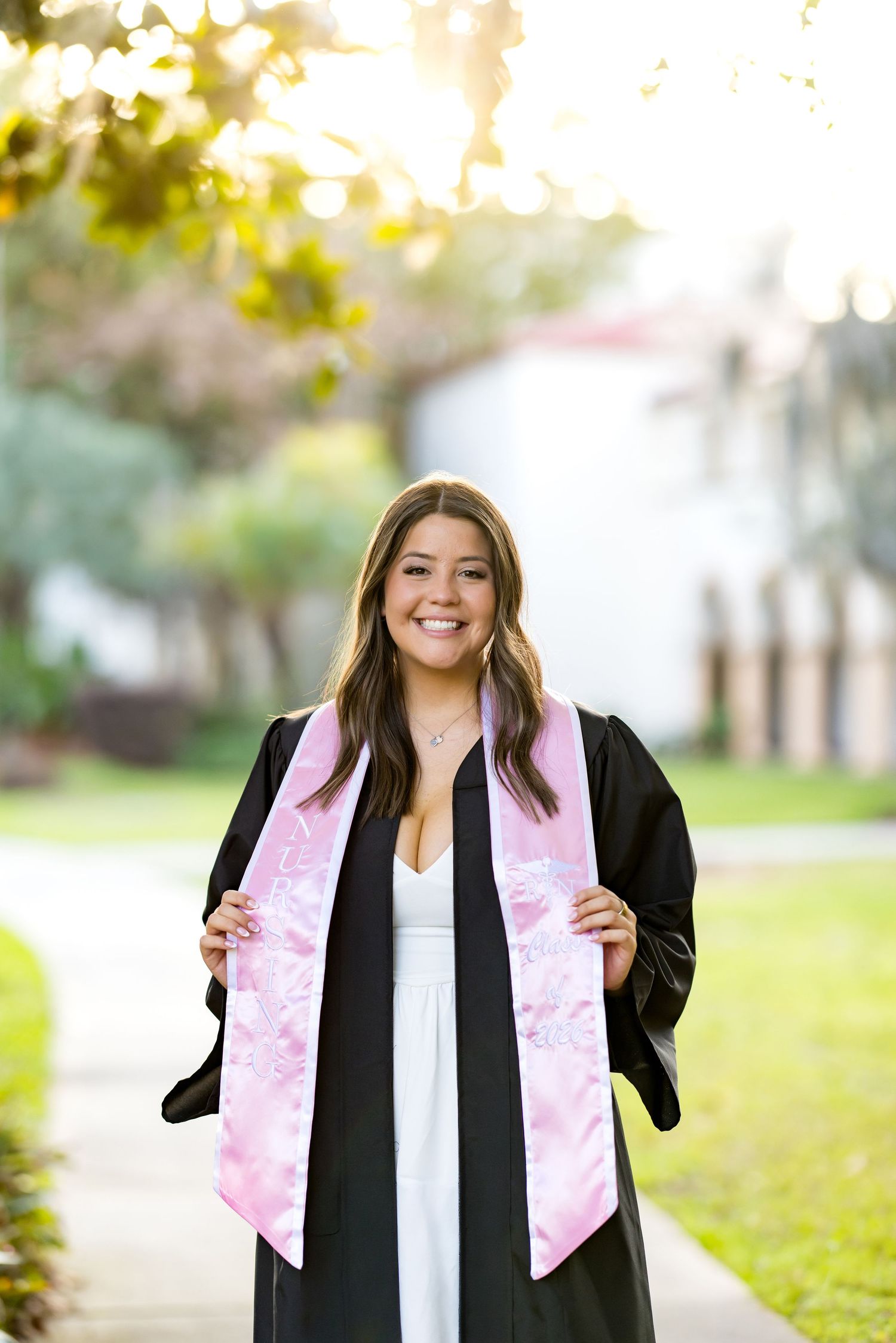 Smiling graduate in black gown and pink floral stole posing outdoors on a sunny campus pathway.