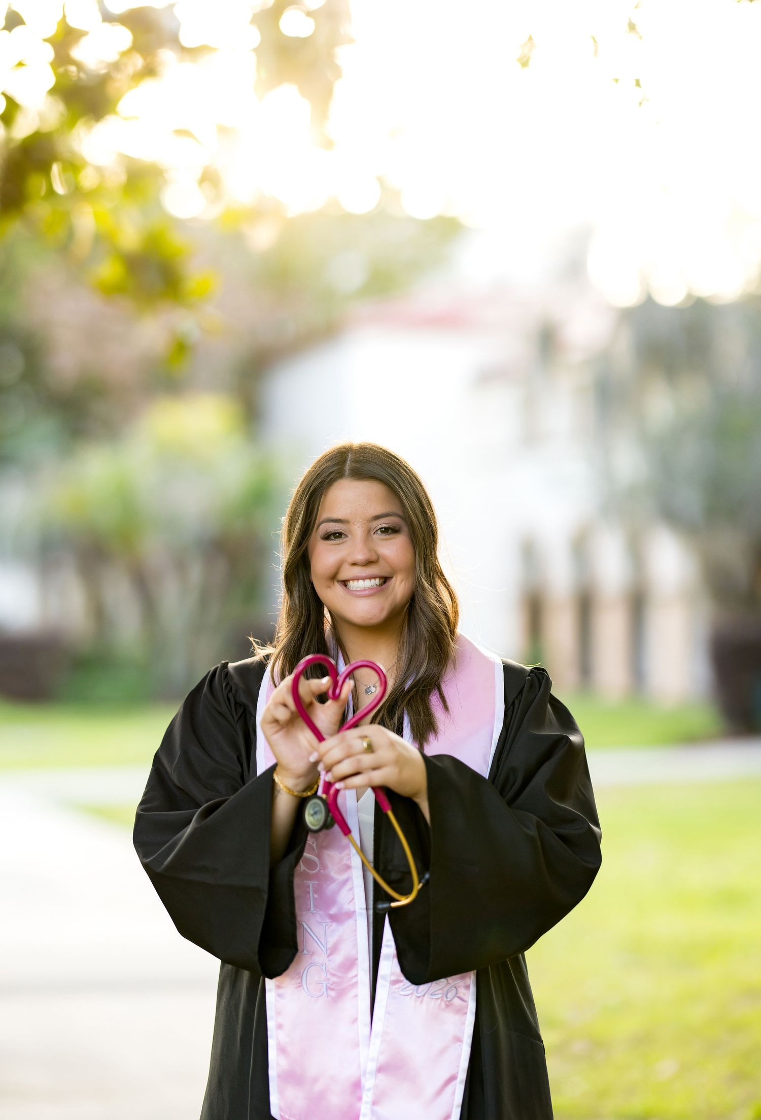 Smiling nursing graduate in black gown and pink stole holds stethoscope shaped as heart outdoors.