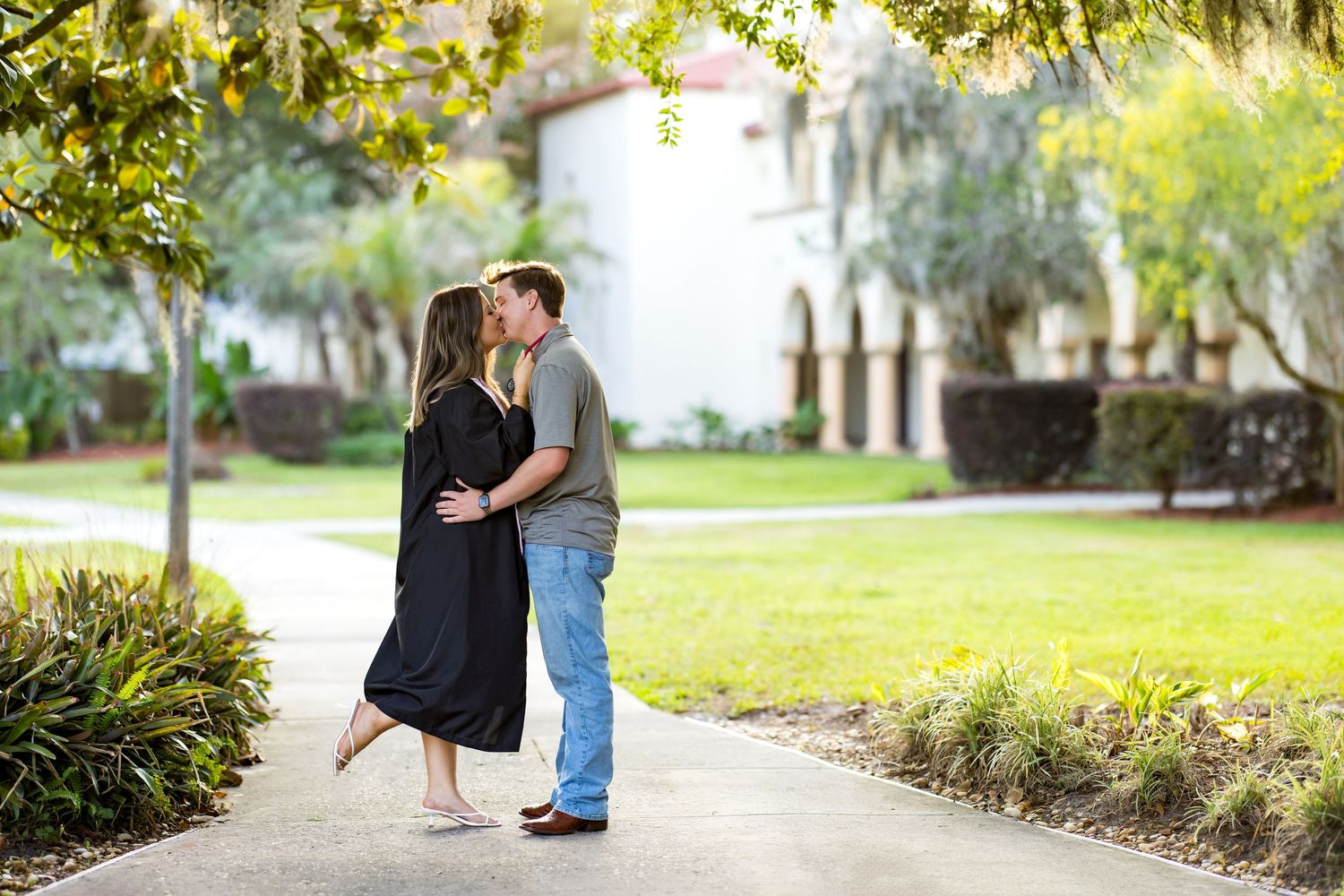 Couple sharing a romantic kiss on a garden path surrounded by lush greenery and a historic building backdrop.