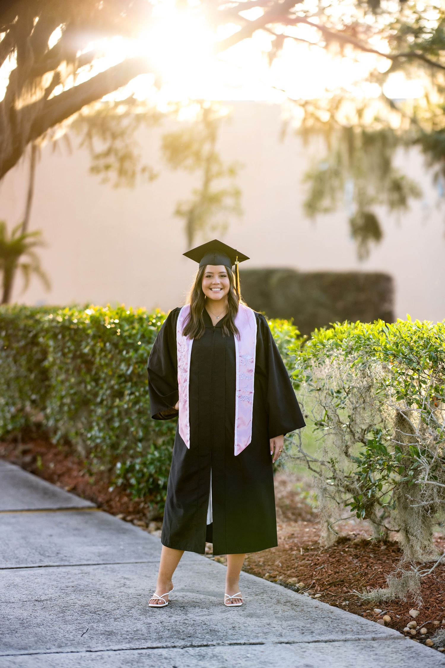 Smiling graduate in black cap and gown with pink sash poses on sunlit garden path surrounded by lush green hedges.