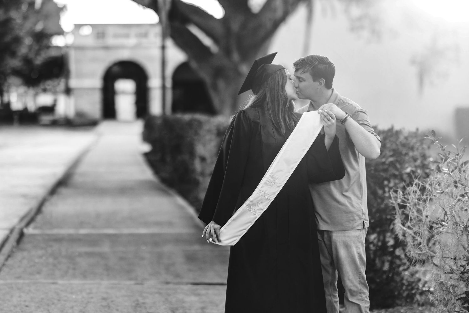 Black and white photo of a graduate in cap and gown embracing a partner outdoors near a historic building.