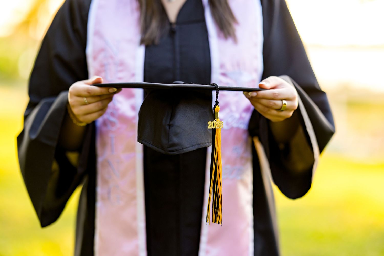 Graduate in black gown holding mortarboard cap with colorful tassel at outdoor graduation ceremony.