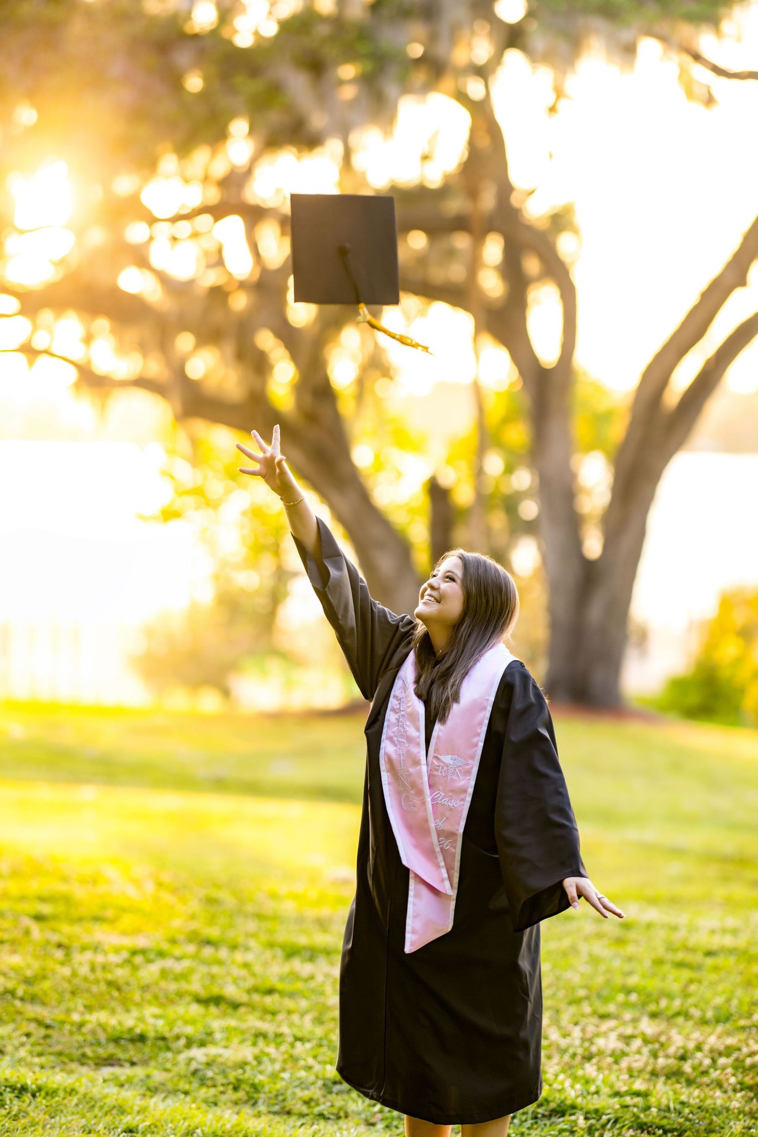 Young woman in graduation gown tossing cap in the air outdoors, celebrating academic achievement at golden hour.