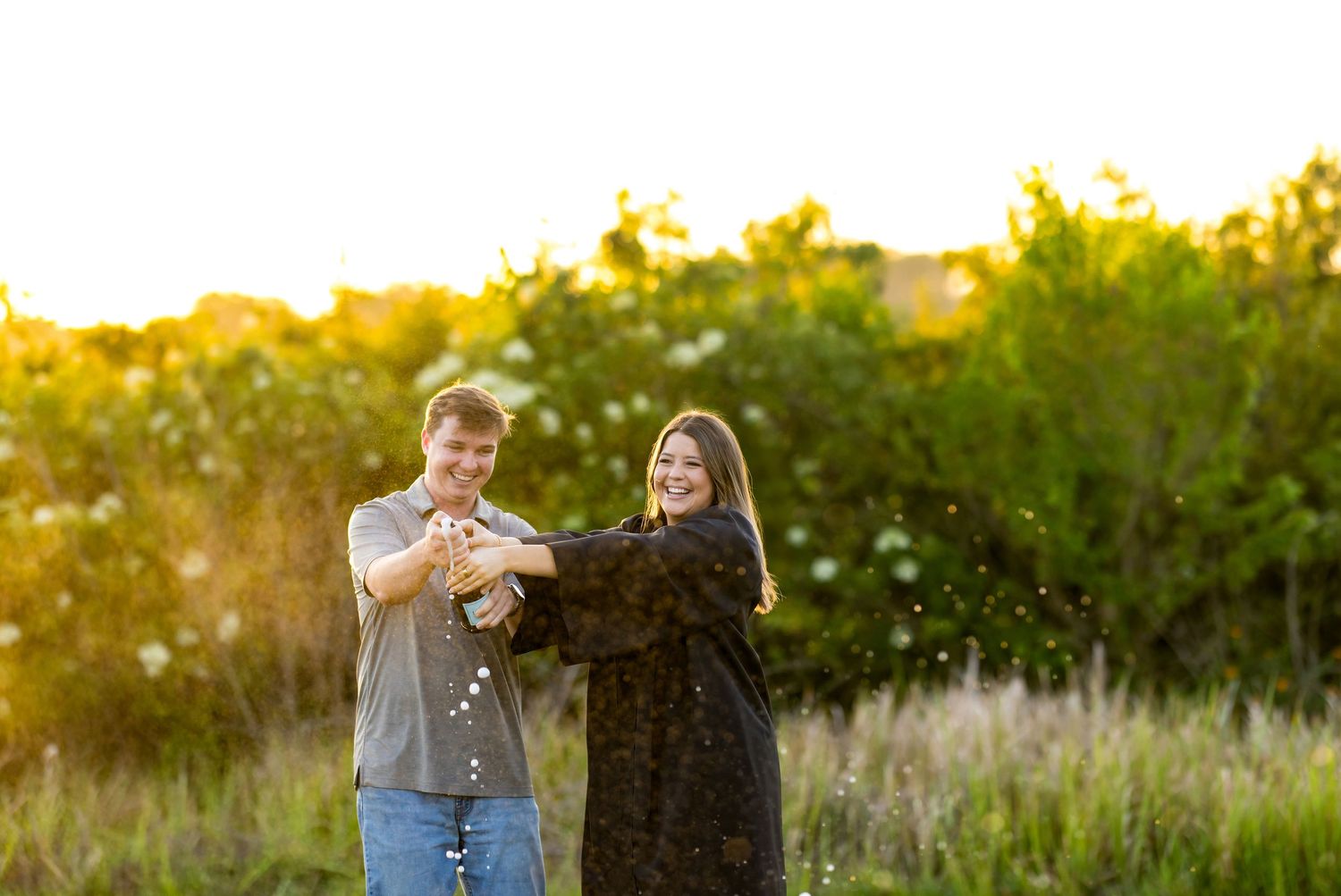Couple popping champagne bottle together outdoors in golden sunset light with lush green trees in background.