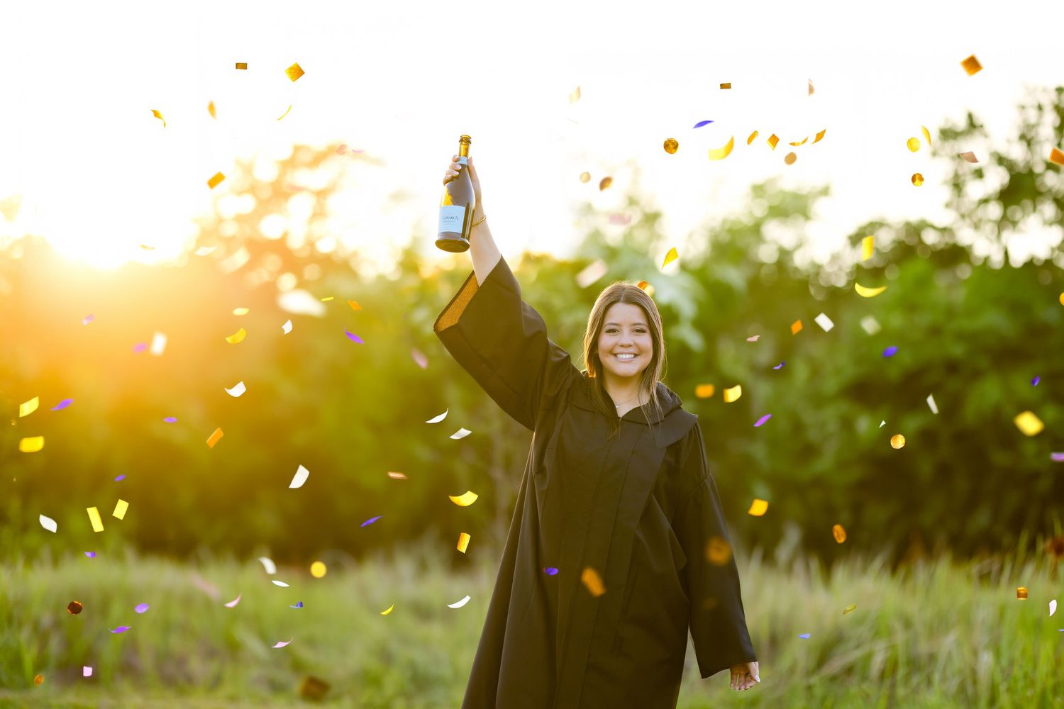 Smiling graduate in black gown holds diploma aloft amid golden confetti in a sunlit outdoor setting.