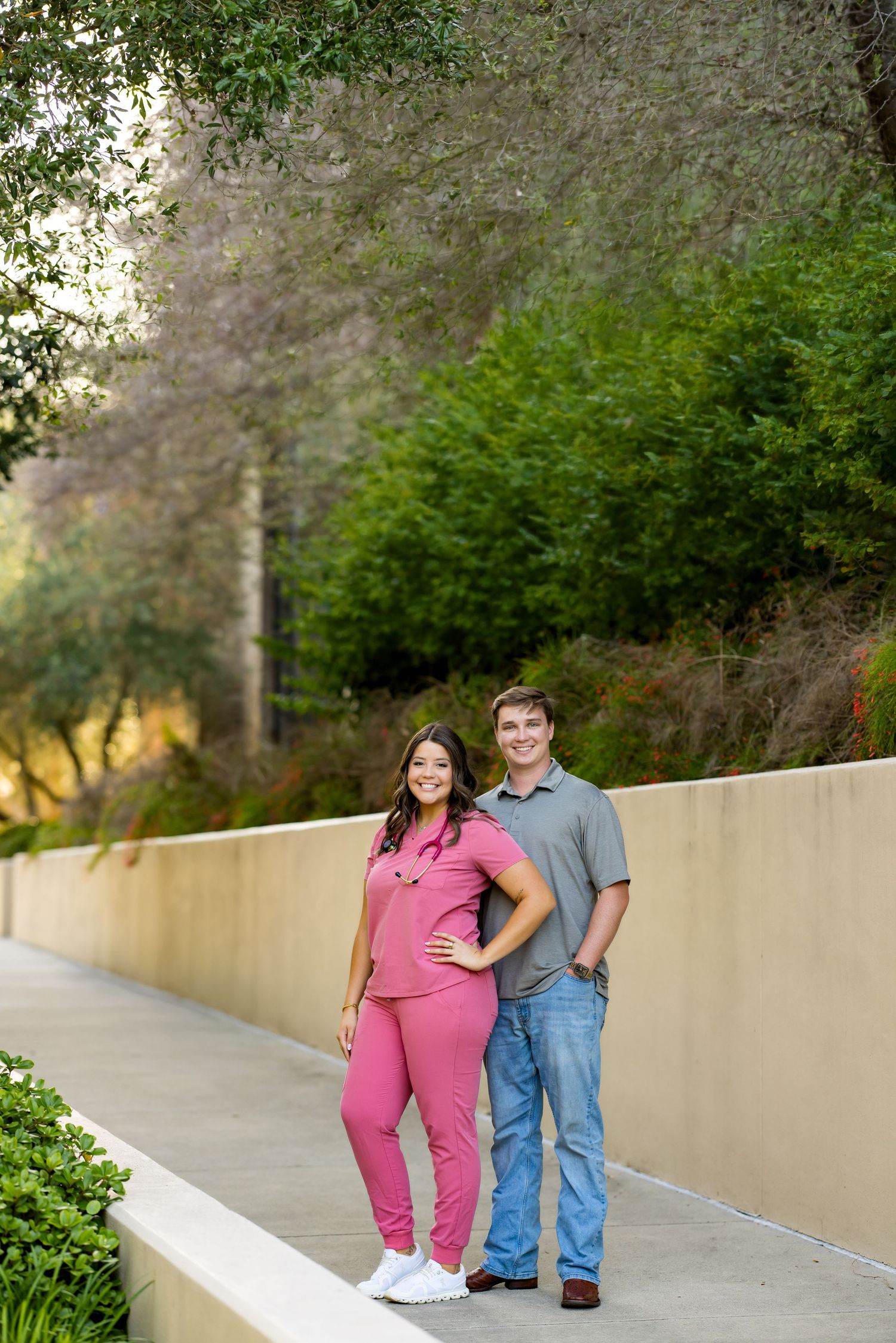 A couple smiling outdoors, woman in pink scrubs and man in gray shirt and jeans, standing against a tan wall.