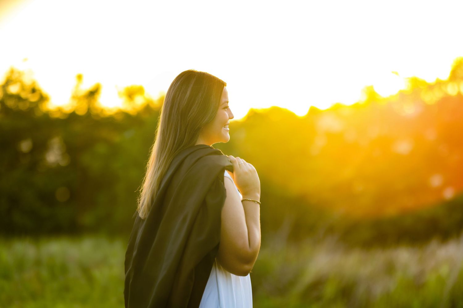 Woman in white dress with dark shawl standing in golden sunset light in a green field.