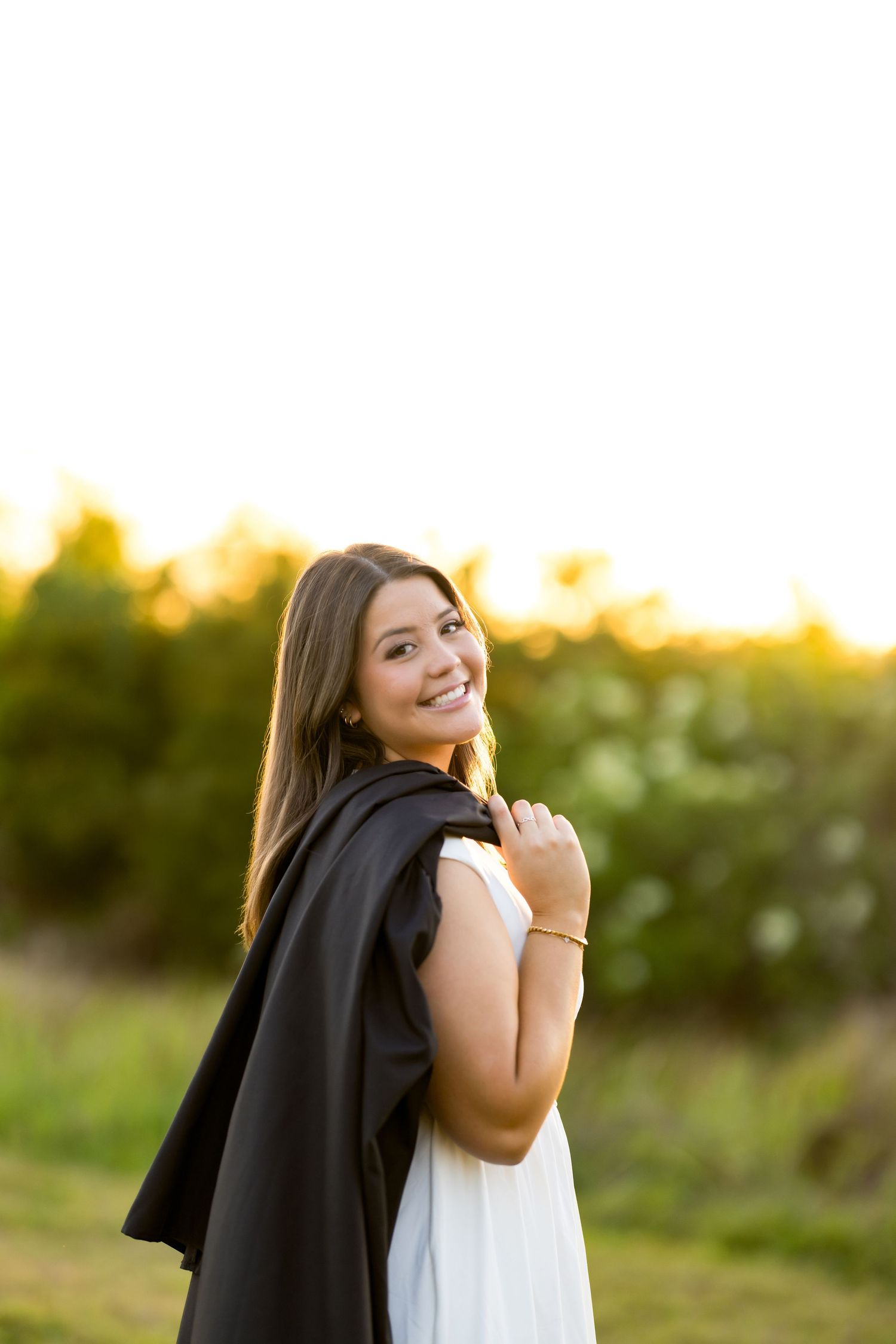 Smiling young woman in white dress holding black graduation gown outdoors at golden hour.