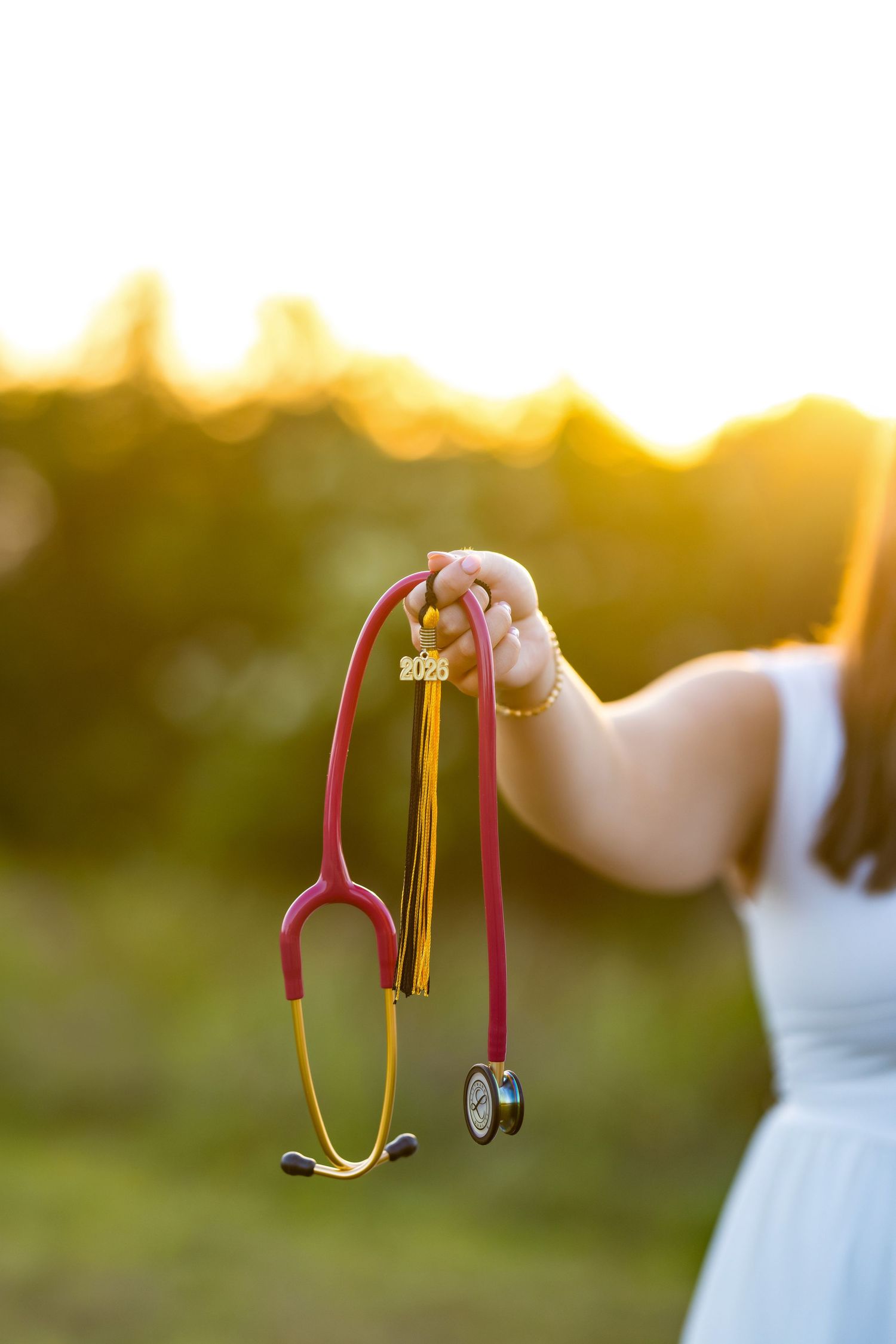 Medical graduate holds stethoscope and graduation tassel outdoors at golden sunset, celebrating healthcare degree achievement.