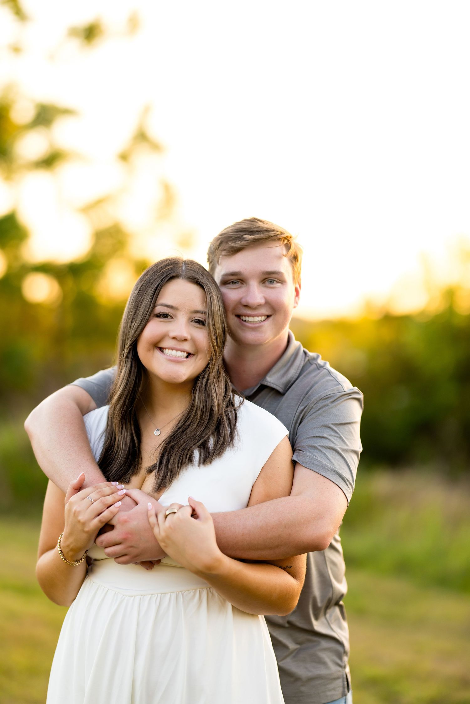 A smiling couple embraces outdoors at golden hour, woman in white dress, man in gray polo, with green trees behind.