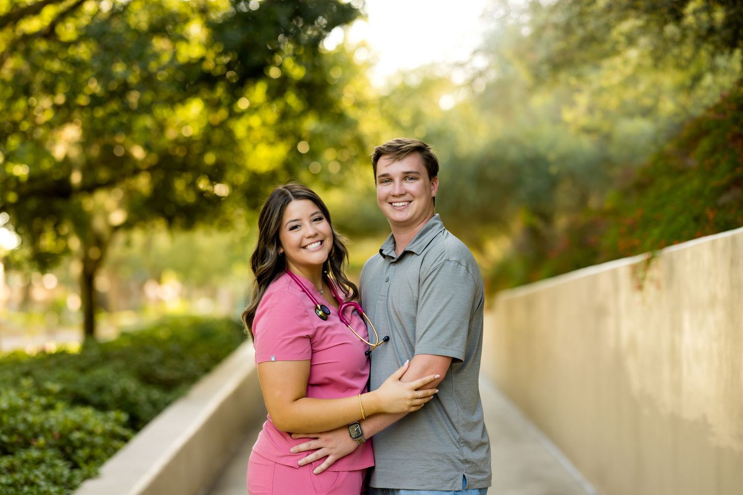 A smiling couple poses outdoors, woman in pink dress and man in gray shirt, embracing on a tree-lined walkway.