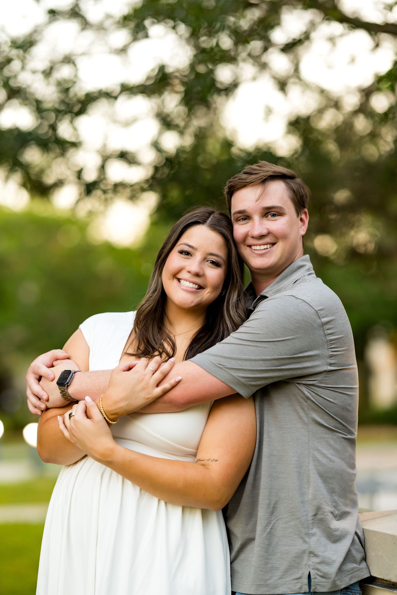 A smiling couple embraces outdoors, woman in white dress, man in gray polo, with lush green trees in background.